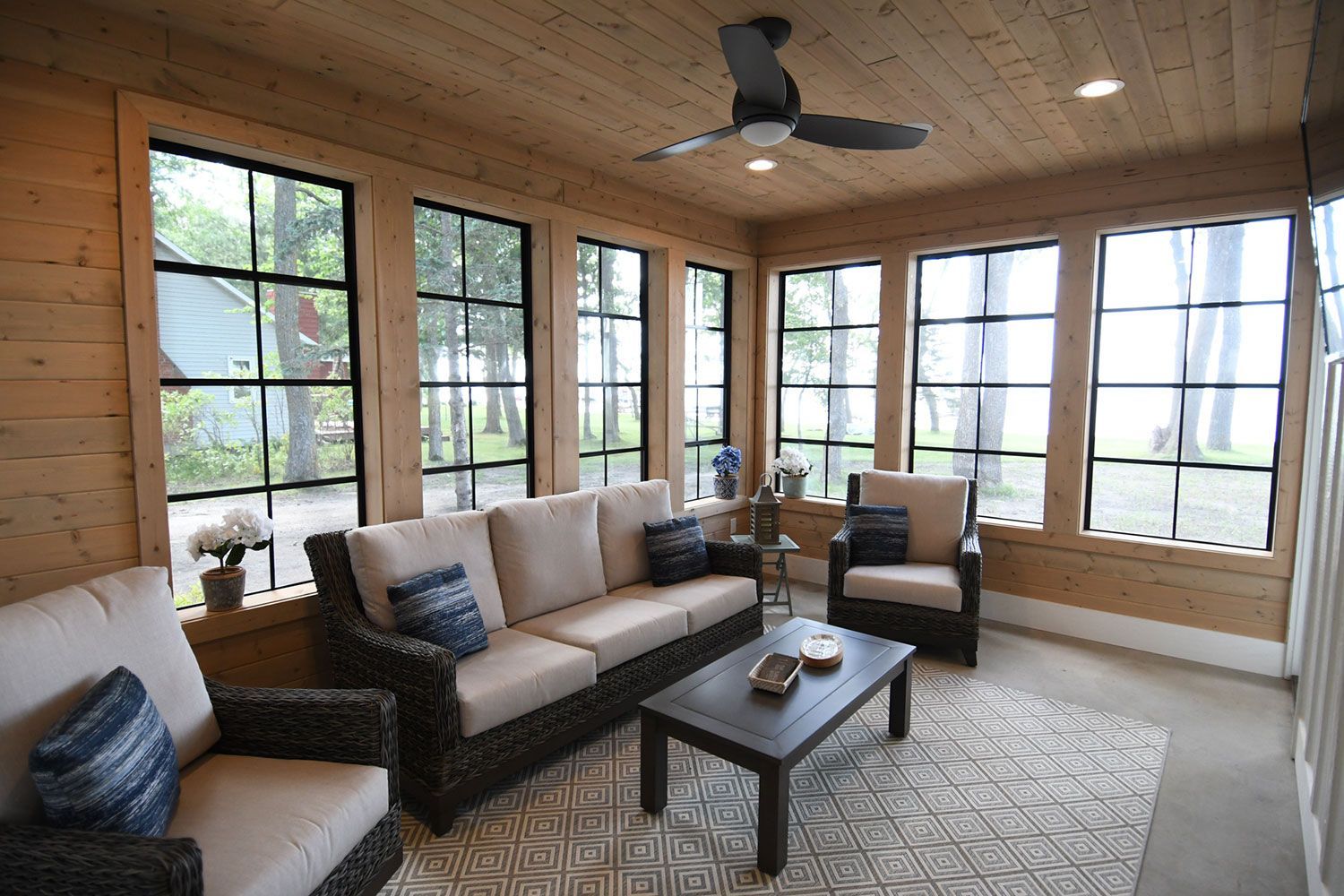 Sunroom with beige sofa, chairs, dark coffee table, rug, and windows overlooking trees.