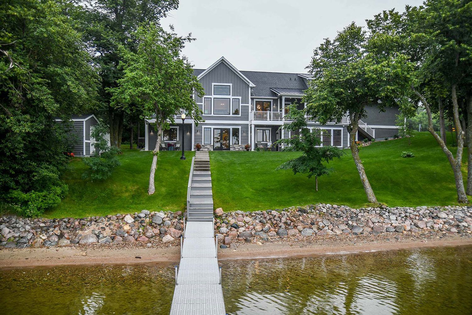 A large gray house by a lake with a wooden dock, surrounded by green grass and trees.