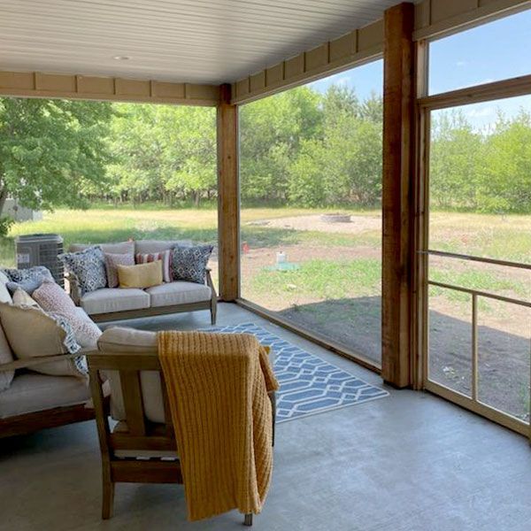Screened porch with wooden furniture, a blue rug, and a yellow blanket, overlooking a grassy yard.