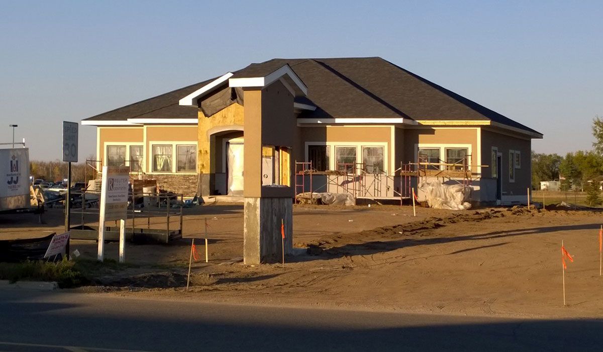 Commercial building under construction with tan siding, gray roof, and stone accents on a sunny day.