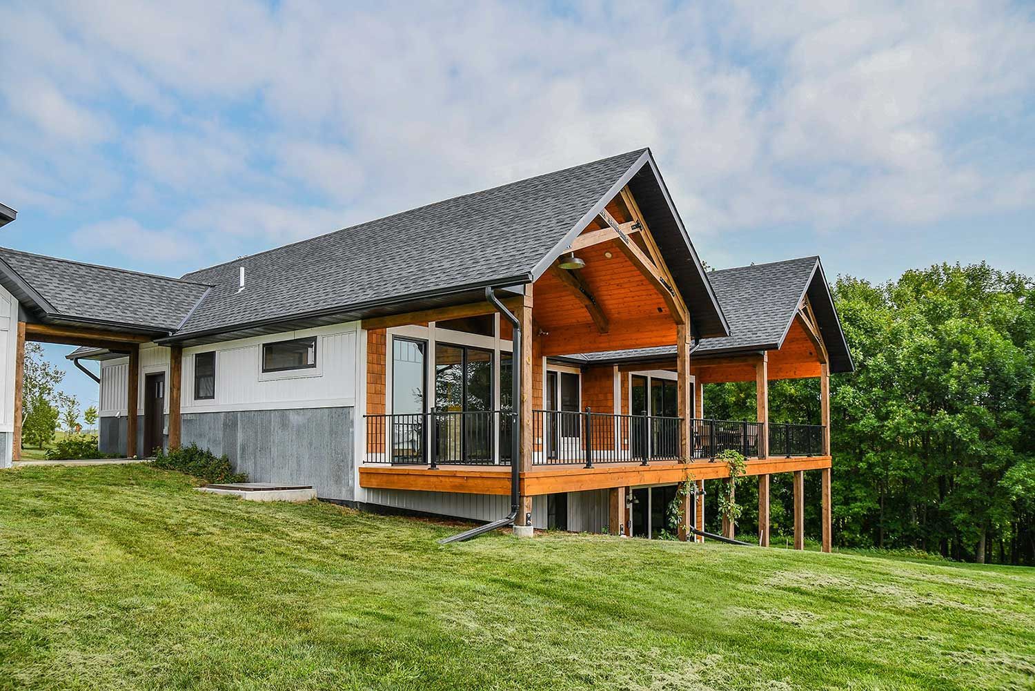 A modern house with a wooden deck and dark roof sits on a grassy hill under a blue sky.