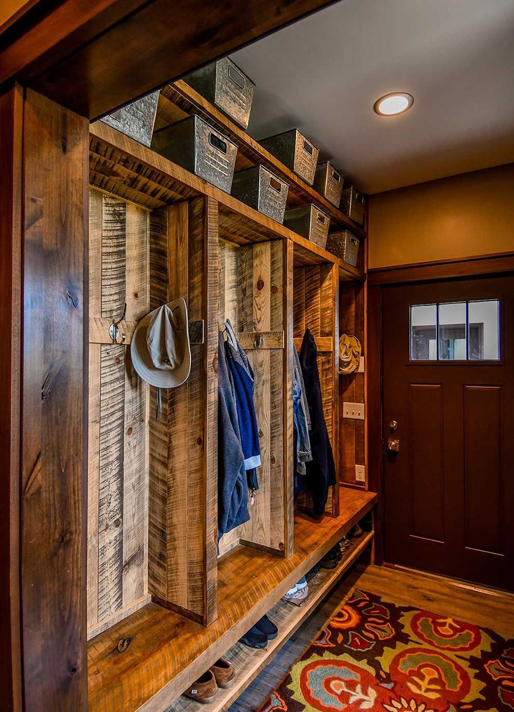 Rustic wooden entryway with coat hooks, storage baskets, and a bench. A hat and coats hang. Door to the right.