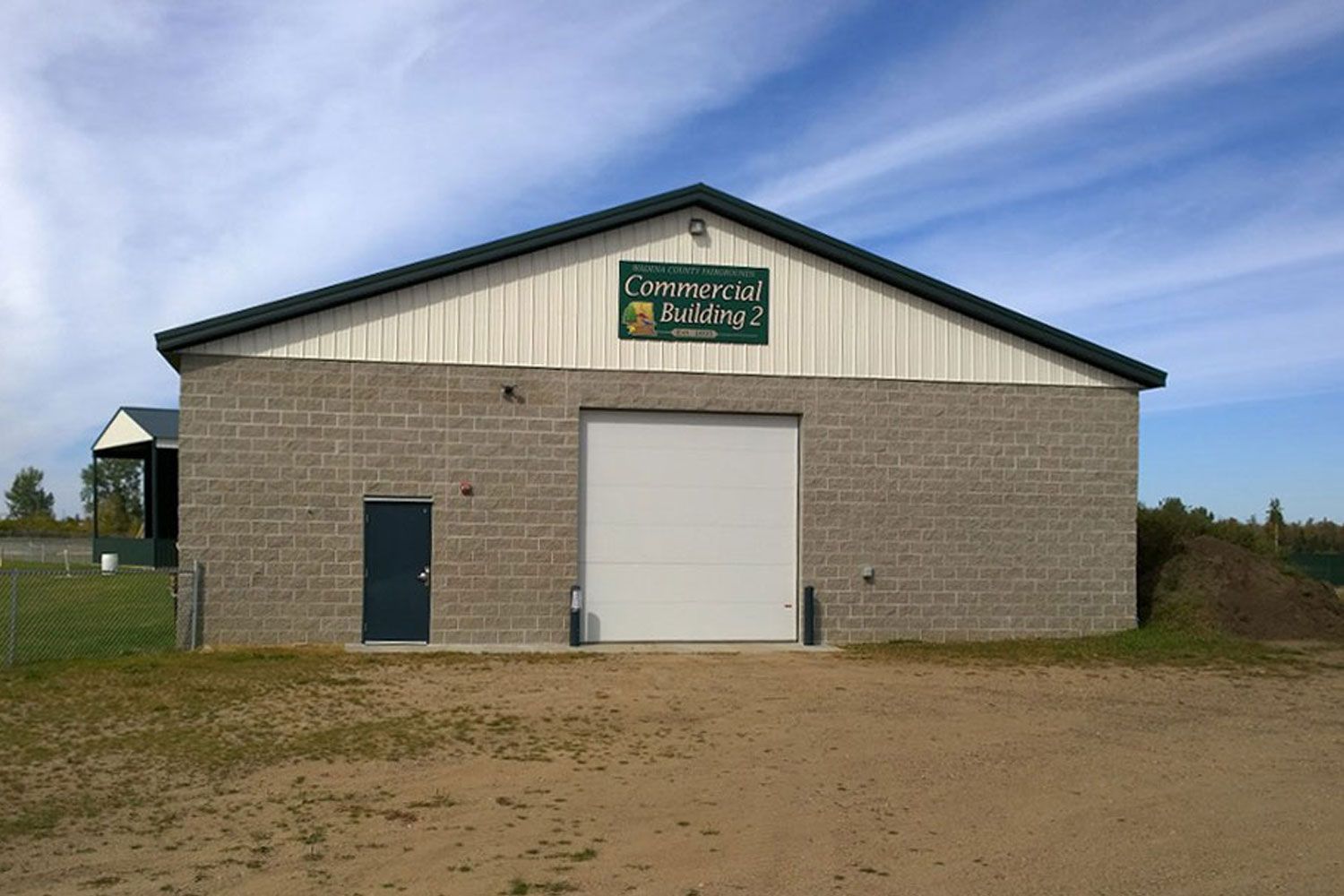 Gray brick building with a large garage door, small door, and a green and white sign; set in a rural area with blue sky.