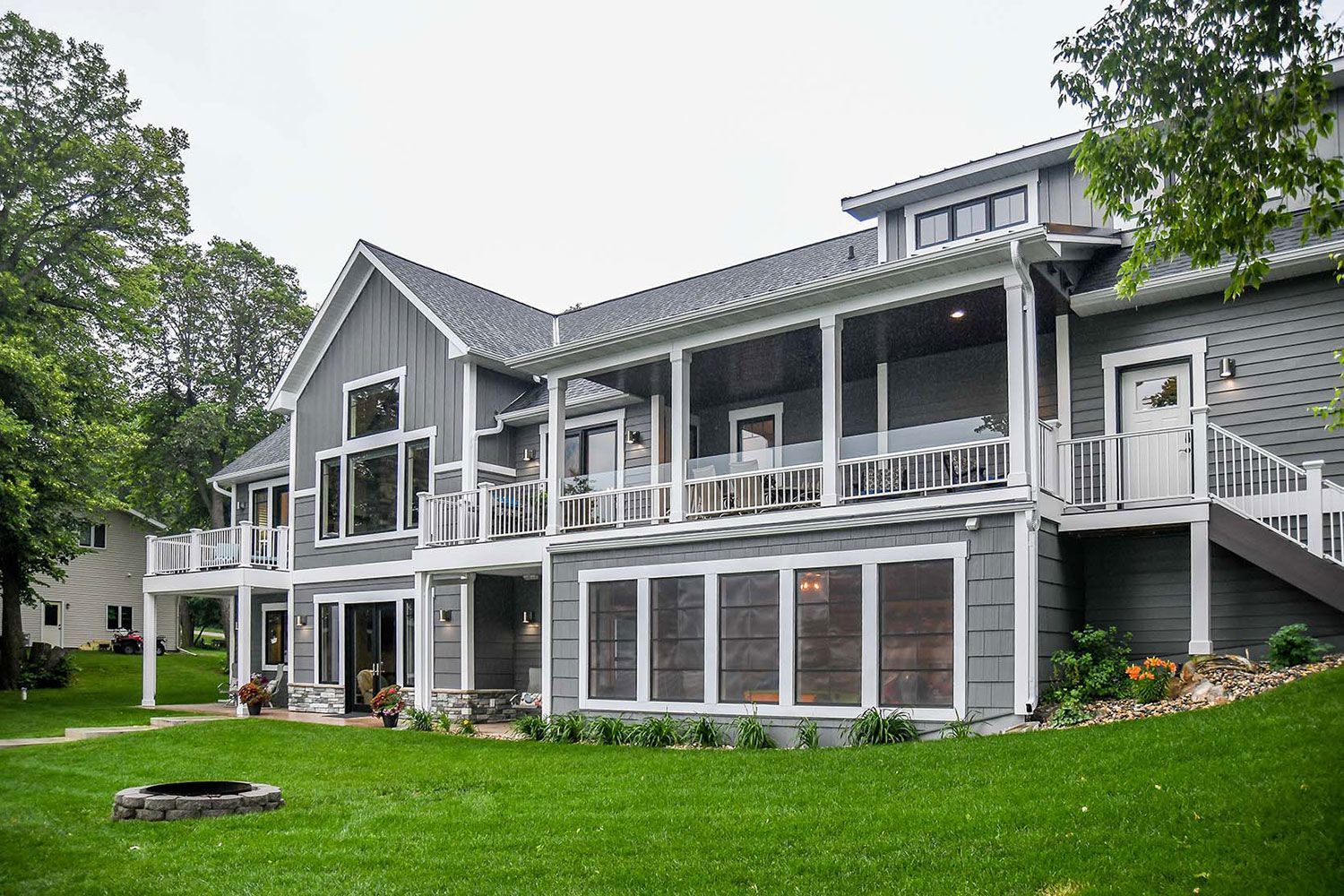 Gray two-story house with porch overlooking a grassy yard and trees. Fire pit in the foreground.