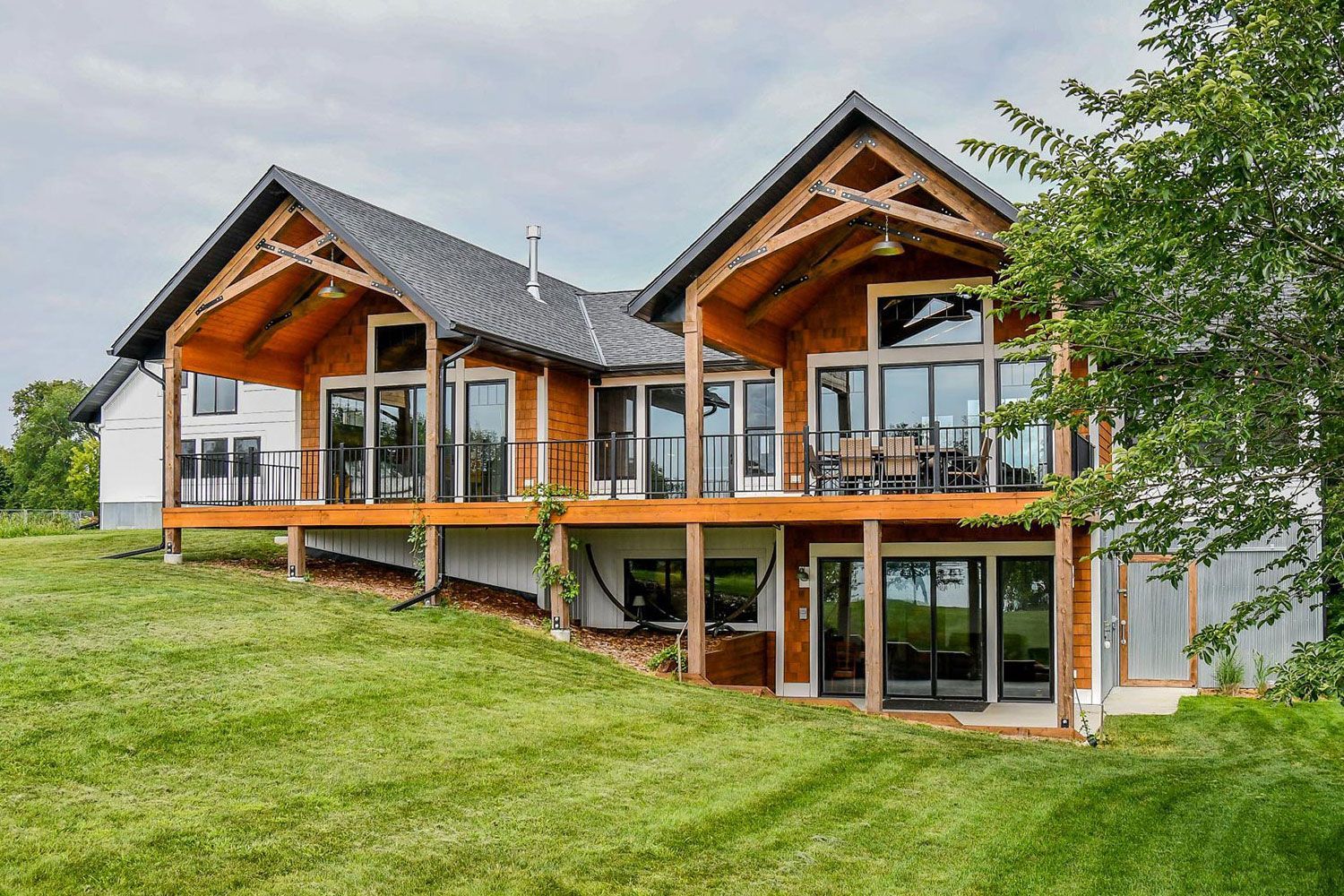 Two-story wooden house with open porch, glass windows, and a sloping green lawn under a cloudy sky.