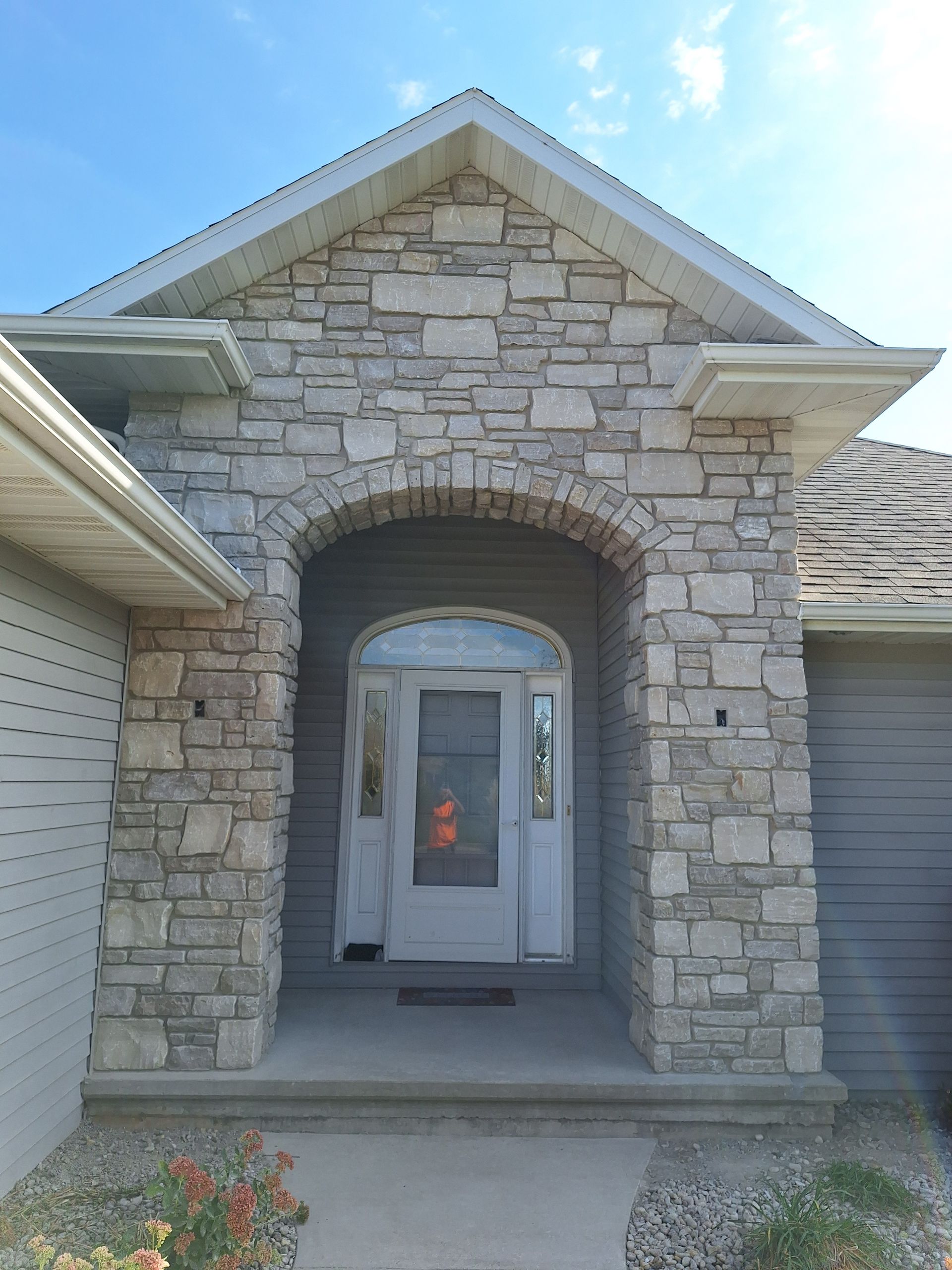 Stone-covered entryway of a house. Arched opening, front door visible. Grey siding and a blue sky.