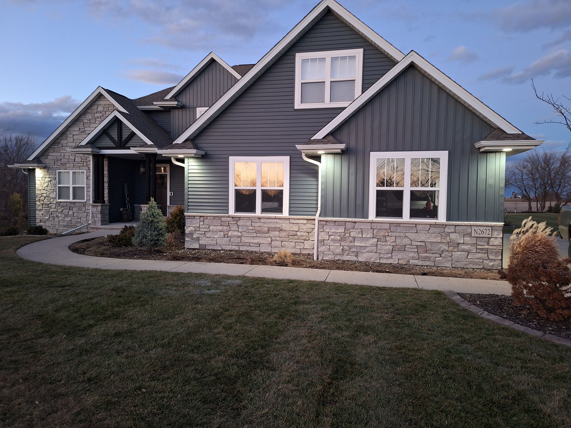 House with blue siding, stone facade, and two gables, lit by exterior lights, dusk setting.