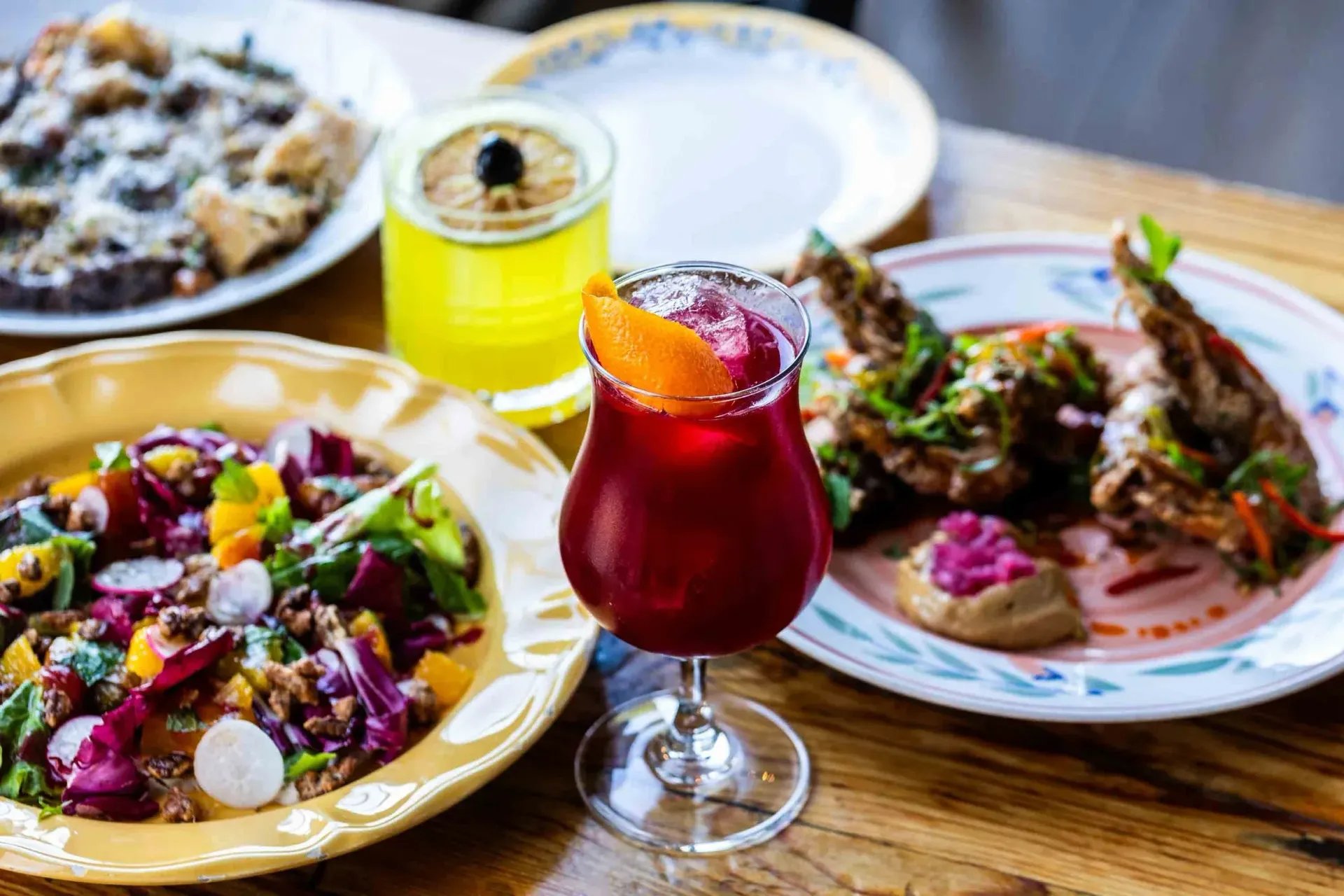 A craft cocktail gleams in the low light at Odd Duck, surrounded by colorful small plates — from fresh market veggies to wood-fired bites — all artfully set on a rustic wooden table in Austin.