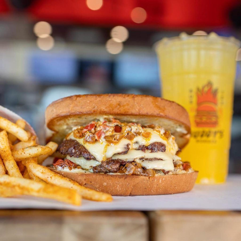 Close-up of a double smashburger with melted cheese, grilled onions, and peppers on toasted bread, served with seasoned fries and a yellow frozen drink at Buddy’s Burgers in Austin.