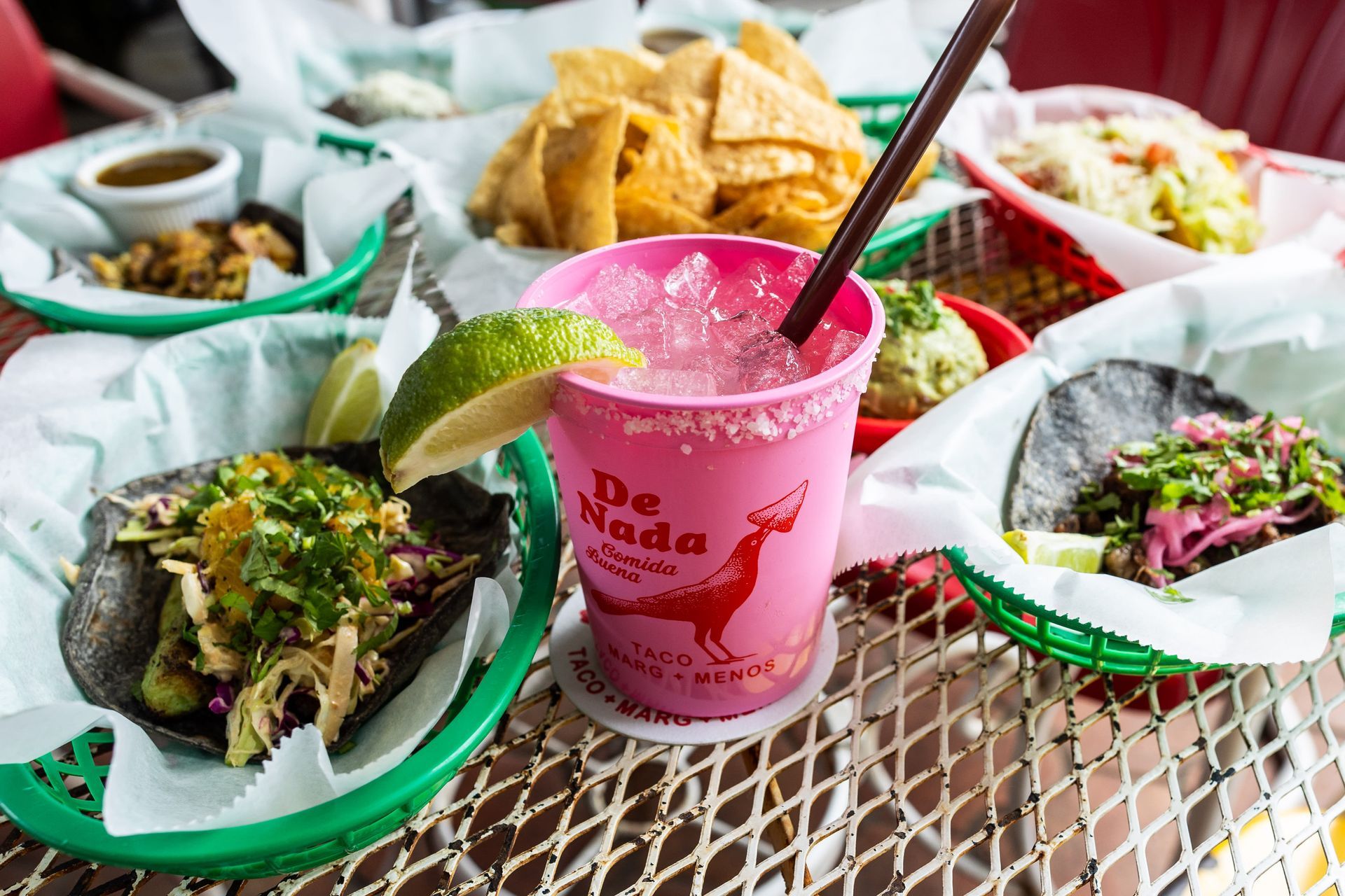 A pink cup with a salted rim and lime wedge filled with a frozen margarita at De Nada Cantina, surrounded by tacos in green baskets, chips, salsa, and guacamole on a metal patio table.