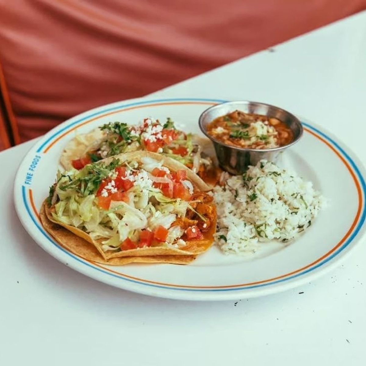 Three crispy tacos topped with lettuce, tomato, and cotija cheese served with cilantro rice and charro beans on a white diner-style plate at Joann’s Fine Foods in Austin.