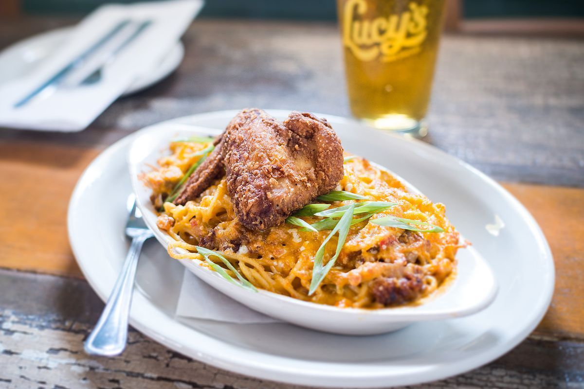 Plate of baked mac and cheese topped with crispy fried chicken wings and green onions, served on a rustic wooden table with a pint of beer in the background at Lucy’s Fried Chicken.