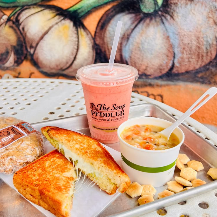 Tray with grilled cheese sandwich, cup of creamy vegetable soup, pink smoothie, and a packaged cookie from The Soup Peddler, set on a metal patio table with a colorful mural background.