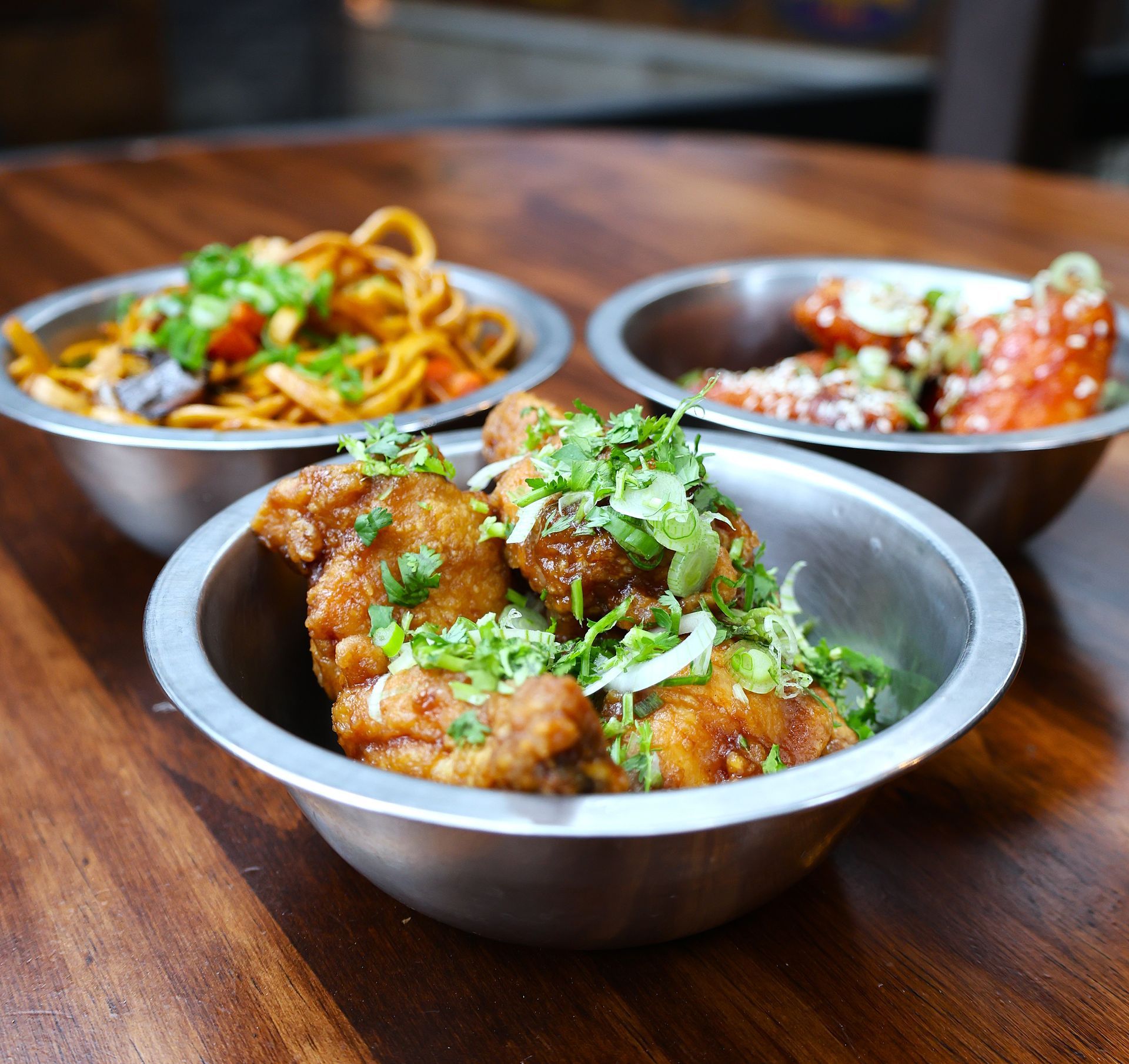 Three metal bowls on a wood table featuring fried chicken wings topped with scallions and cilantro, spicy noodles with vegetables, and saucy wings at Tigers Den in Austin.