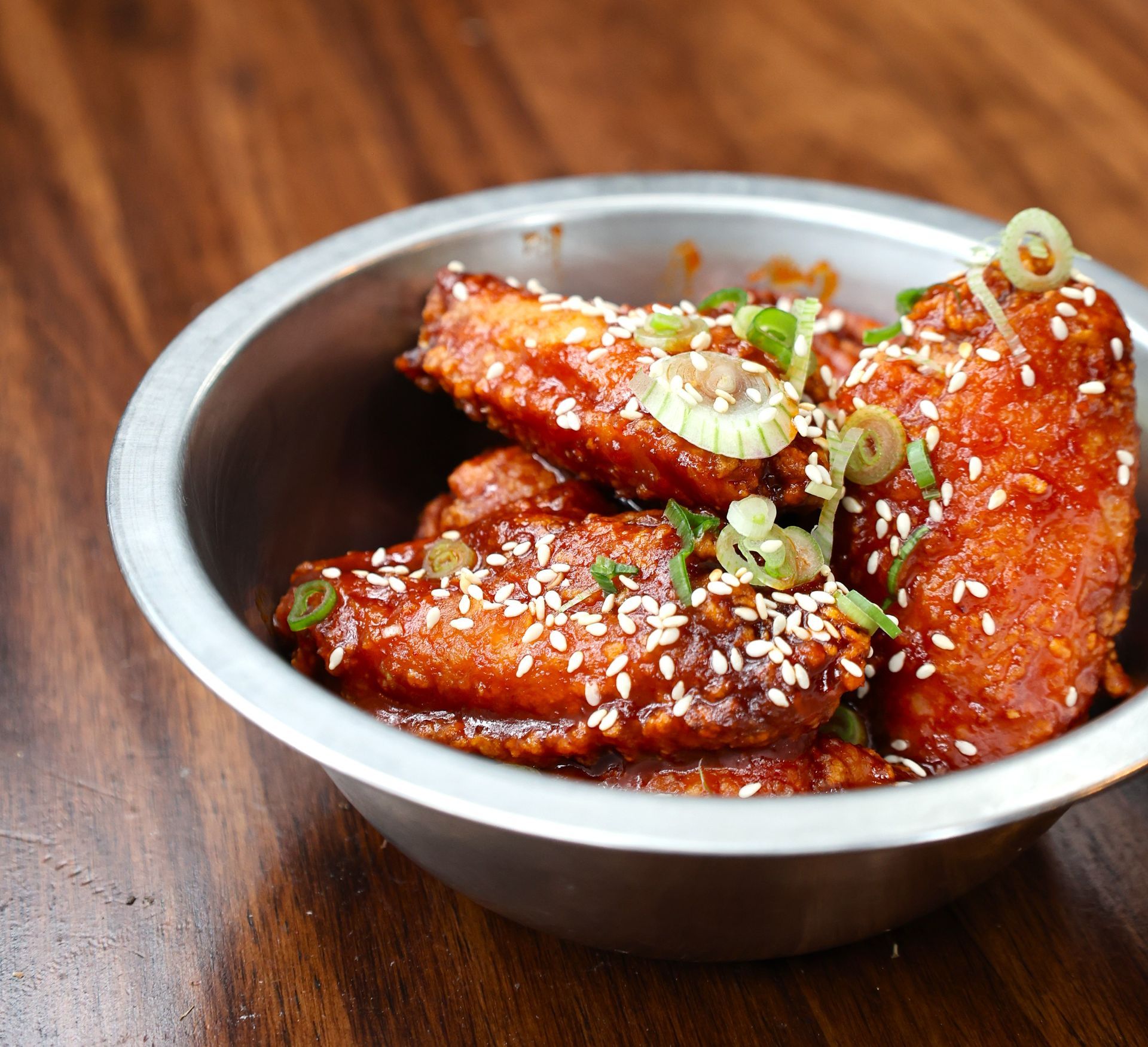 Spicy glazed chicken wings topped with sesame seeds and scallions in a silver bowl on a wooden table at Tigers Den in Austin.