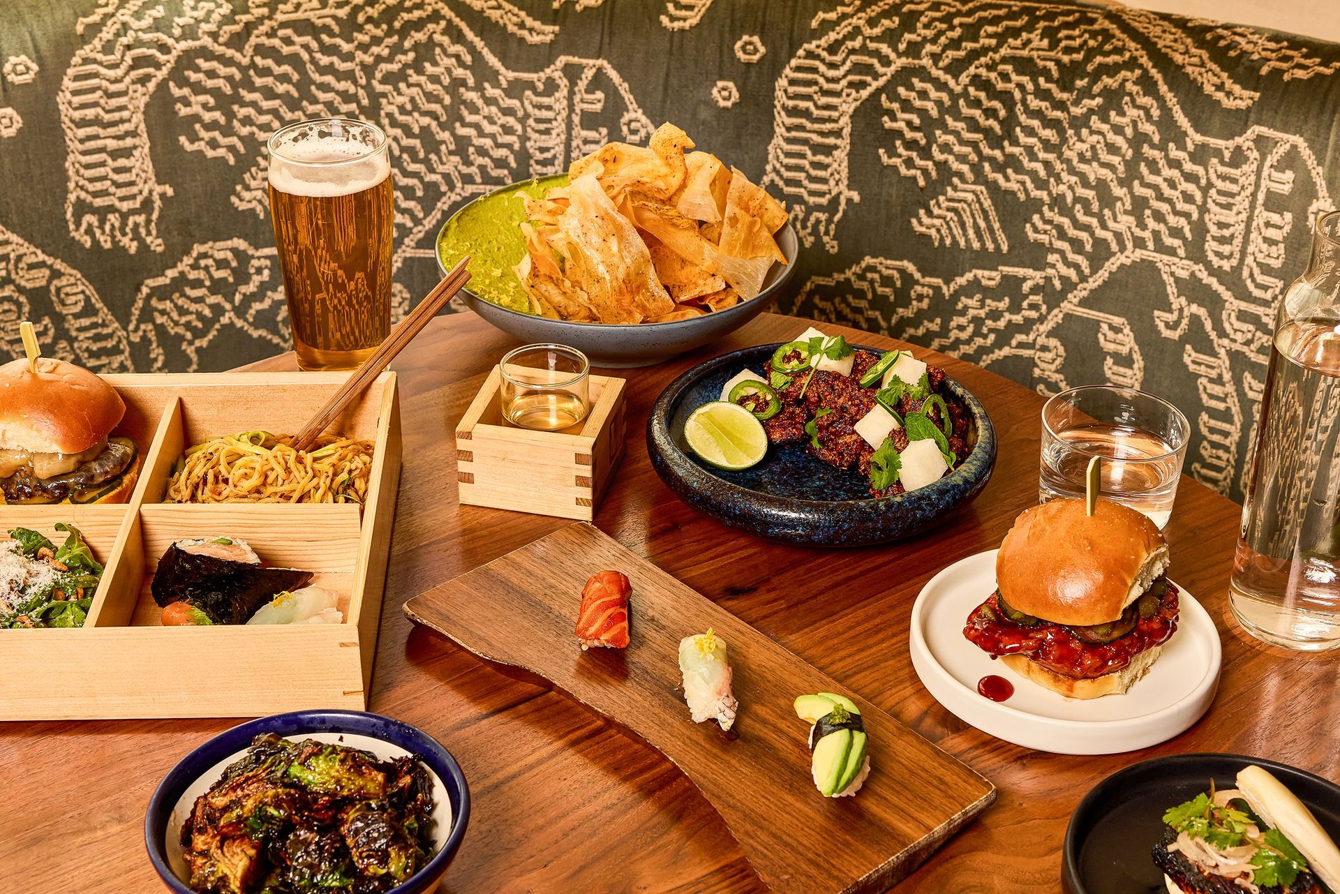 Spread of upscale lunch dishes including sliders, sushi, brussels sprouts, bento-style noodles, chips with green dip, and beer, arranged on a wood table inside Walton’s Fancy & Staple with a patterned booth backdrop.