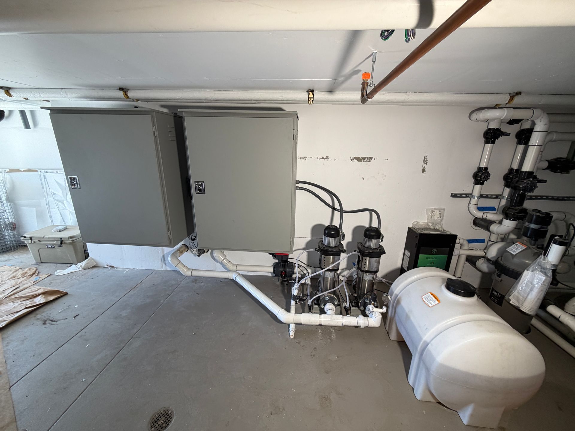 Basement utility room with gray cabinets, pipes, valves, and a white pressure tank on a concrete floor.