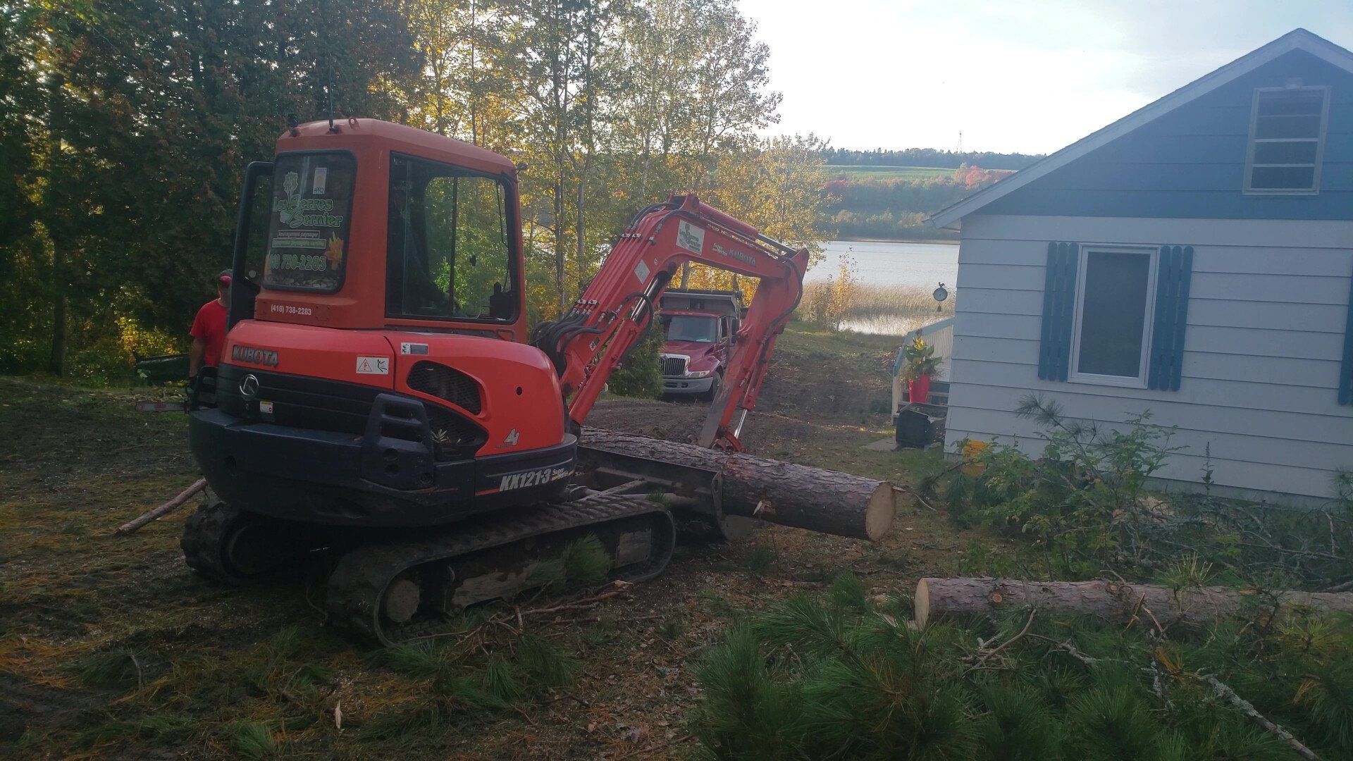 Une excavatrice rouge transporte une bûche devant une maison.