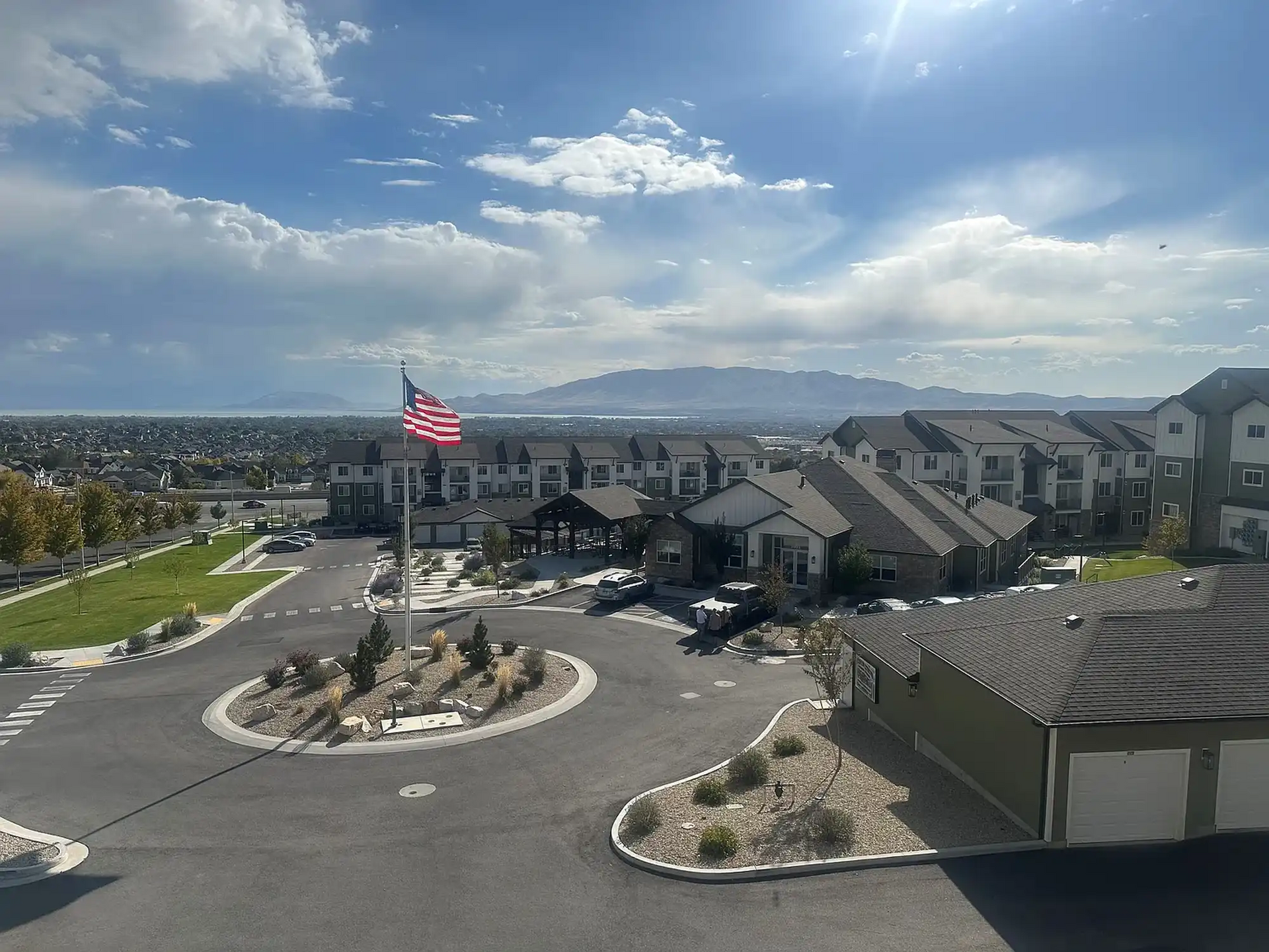 Residential complex with an American flag, circular drive, and mountains in the background under a cloudy sky.