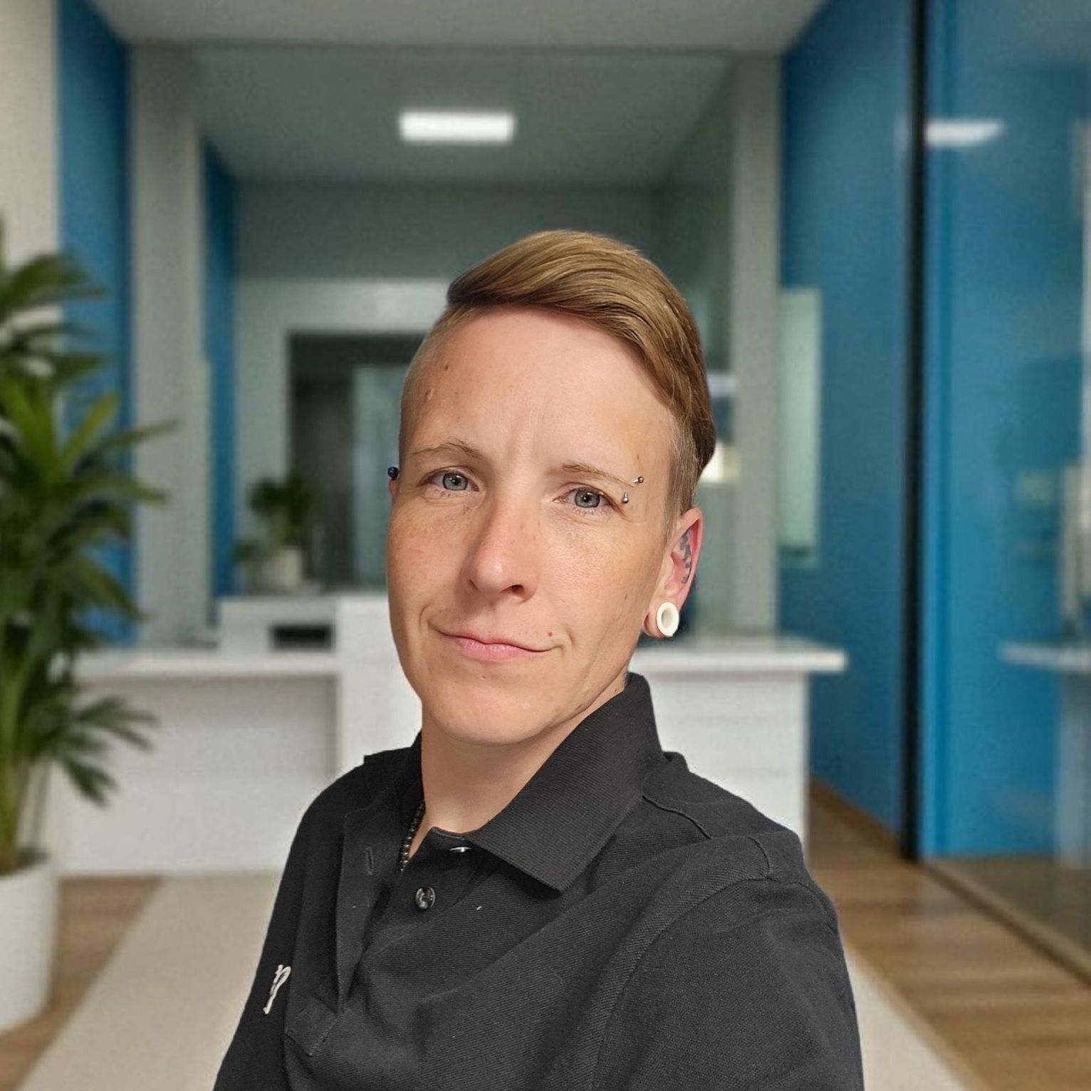 Woman with short, light-brown hair in a black polo shirt, looking at the camera. Office setting with blue walls and greenery.