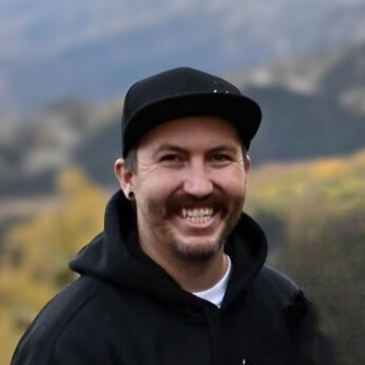 Man with short brown hair and stubble smiling, wearing a black shirt, outdoors in front of a tree.