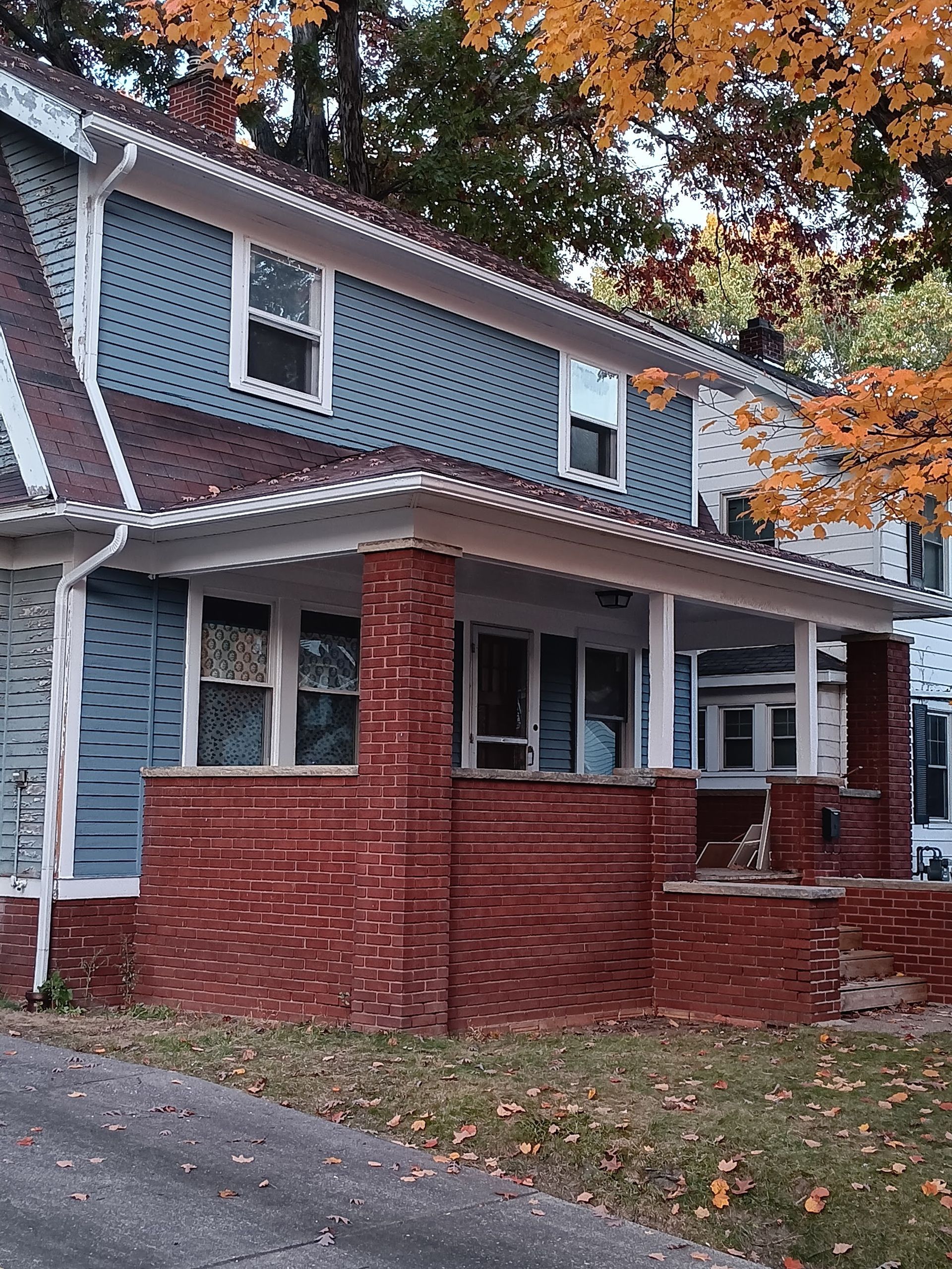 Two-story house with blue siding, red brick porch, and autumn leaves.