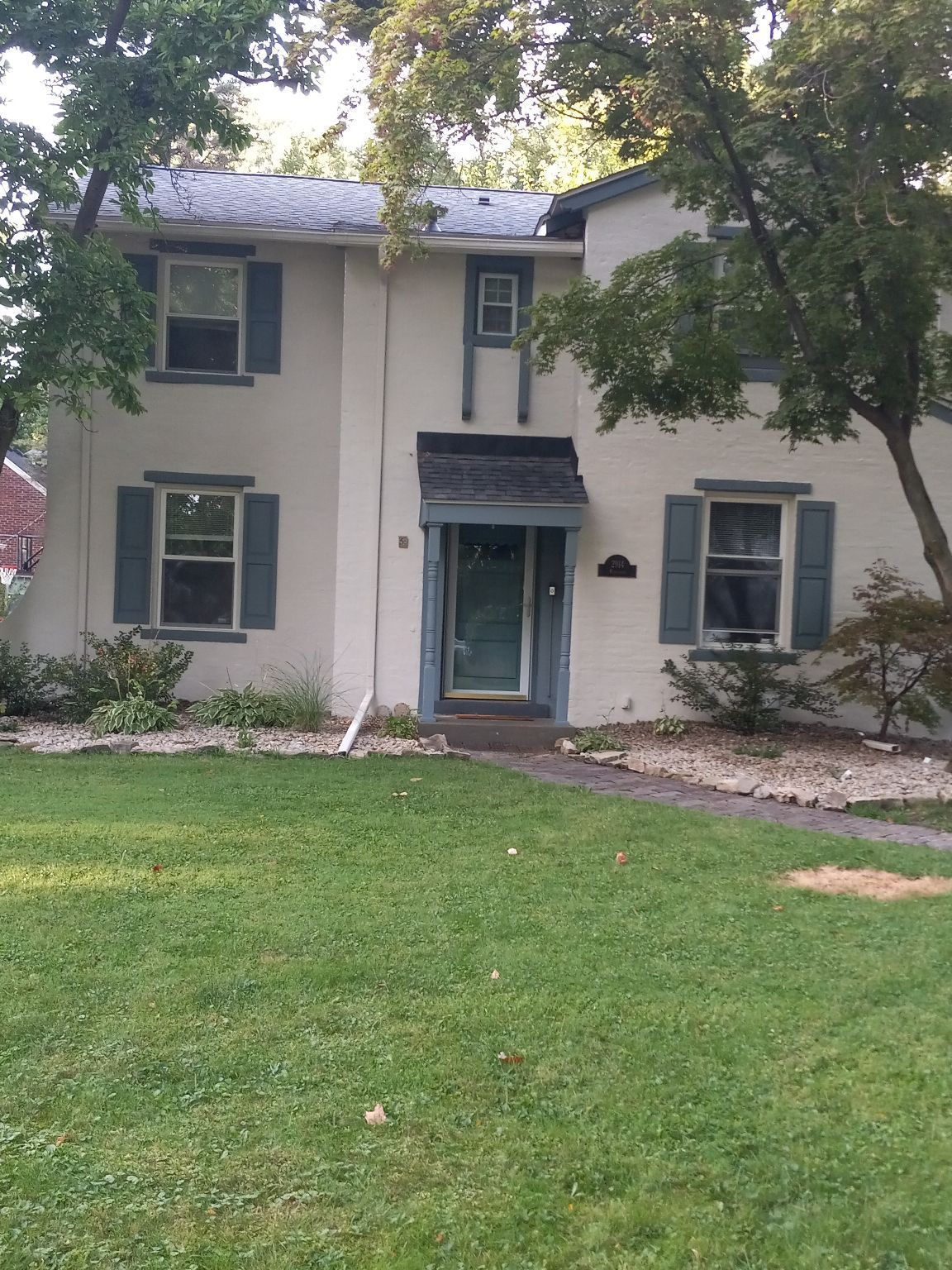 Two-story beige house with blue shutters and door, surrounded by trees and green grass.