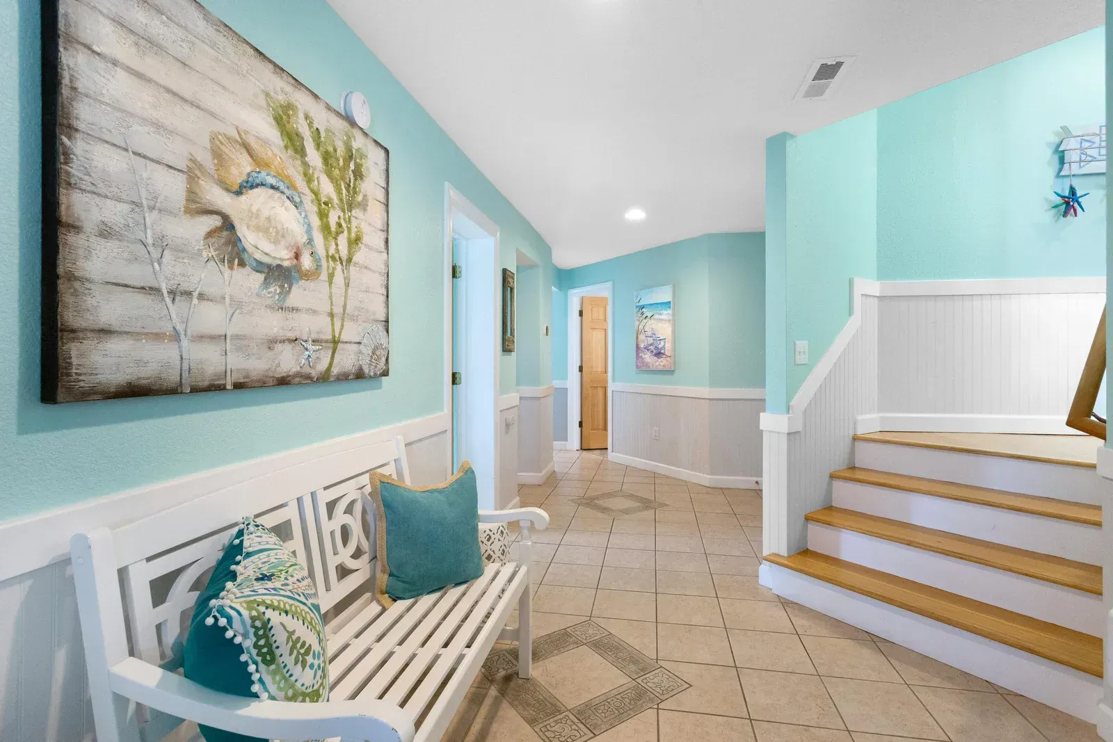Coastal hallway with light blue walls, white trim, bench, artwork, and staircase.