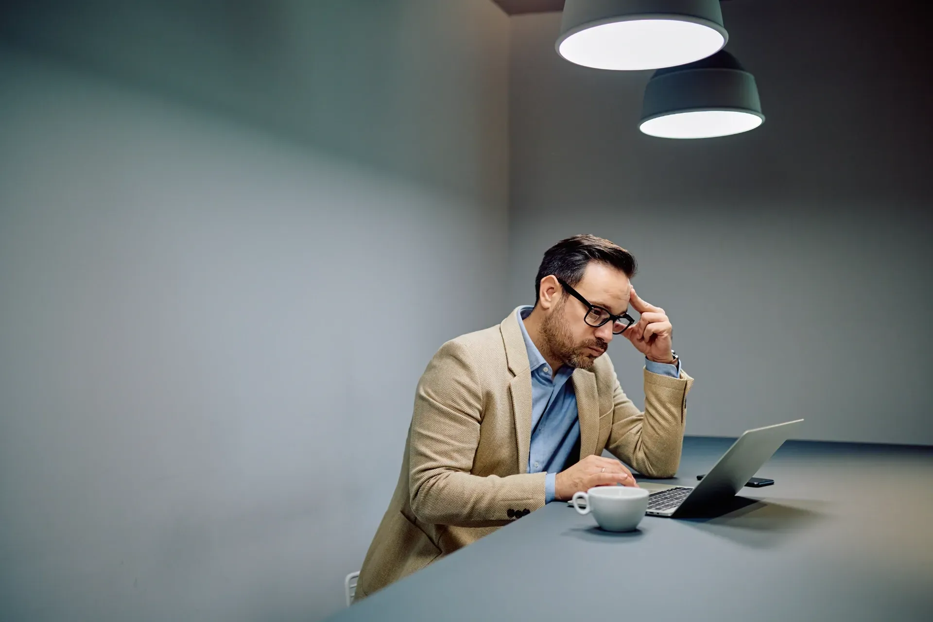 A person in a beige blazer sits at a desk, looking at a laptop with a thoughtful expression, under warm hanging lights.