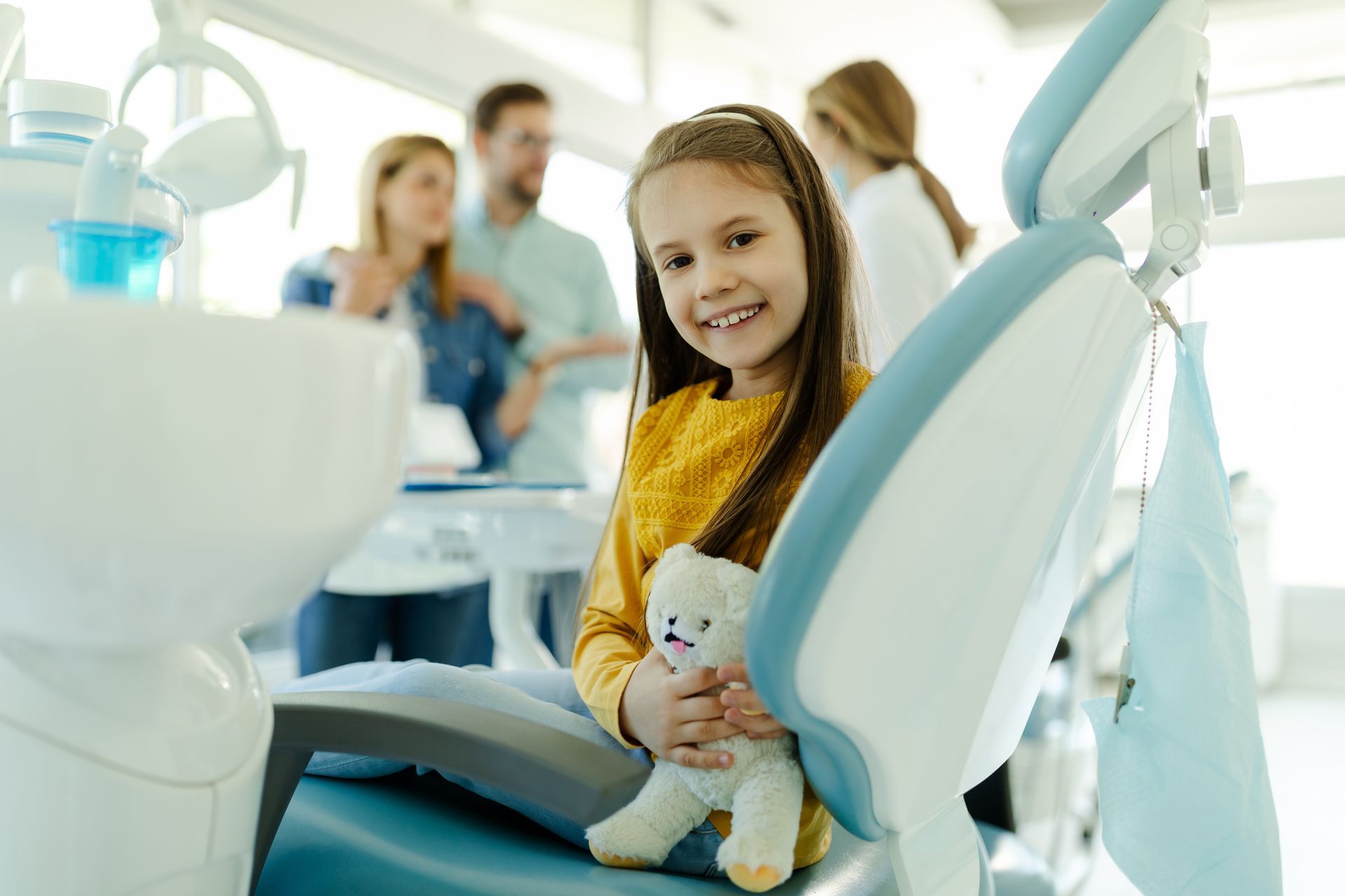 A child with a toy sits in a dental chair as a dentist prepares a check up.