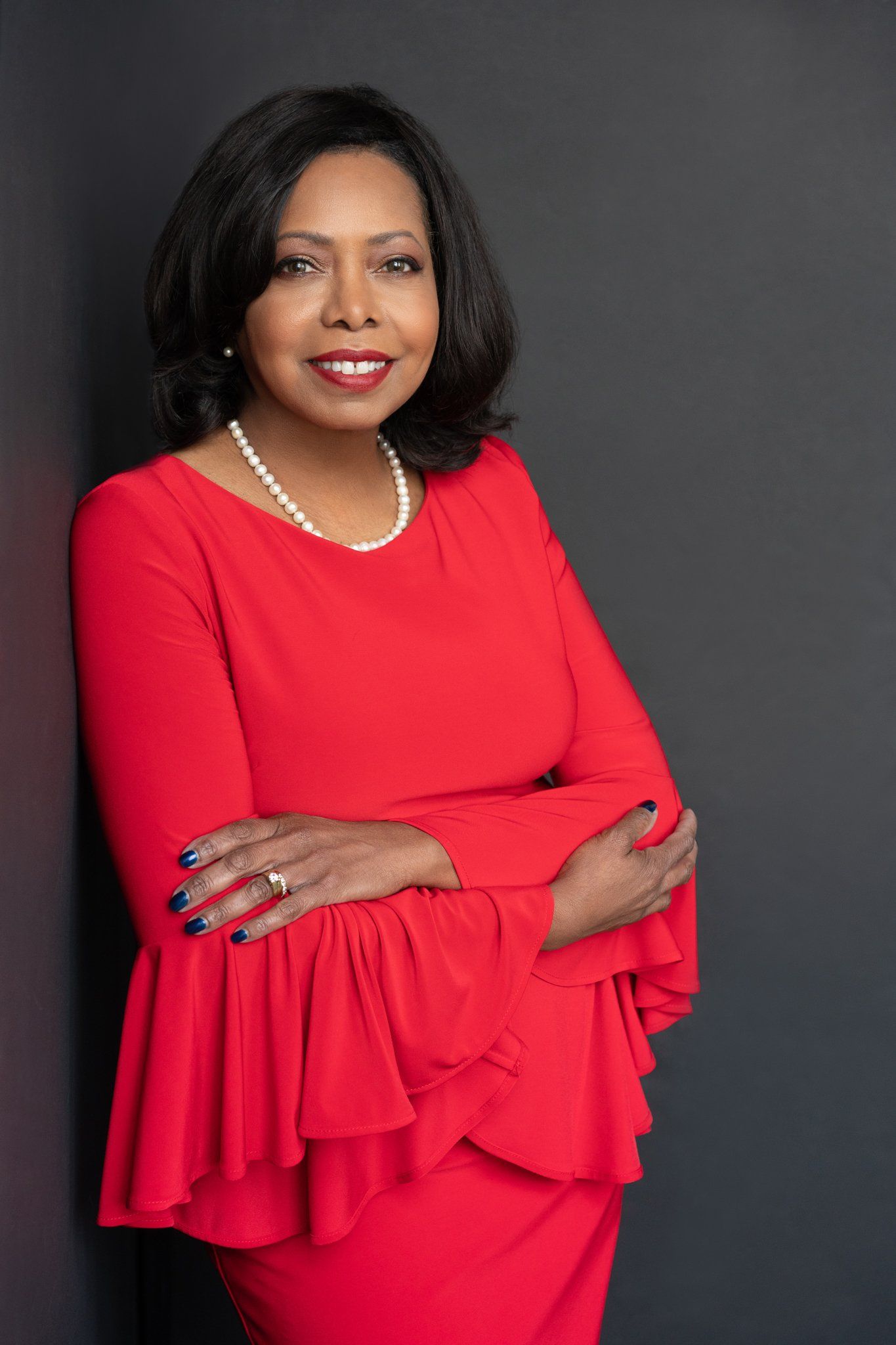 A woman in a red dress and pearls is standing with her arms crossed.