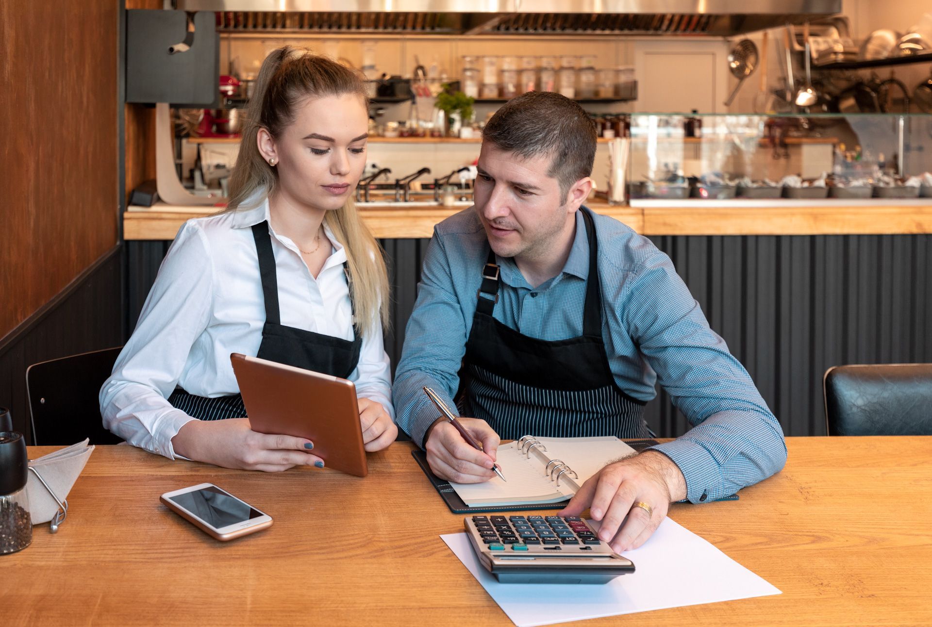 A man and a woman are sitting at a table in a restaurant looking at a tablet and a calculator