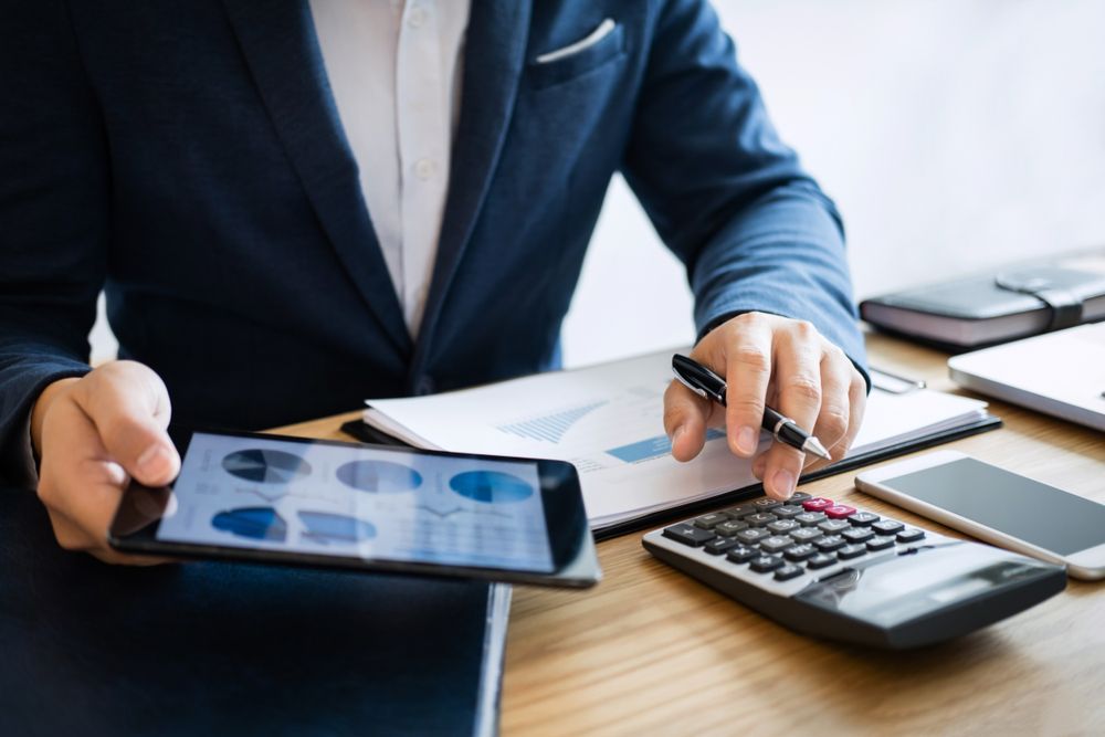 A person in a suit reviews financial data on a tablet while using a calculator at a desk.