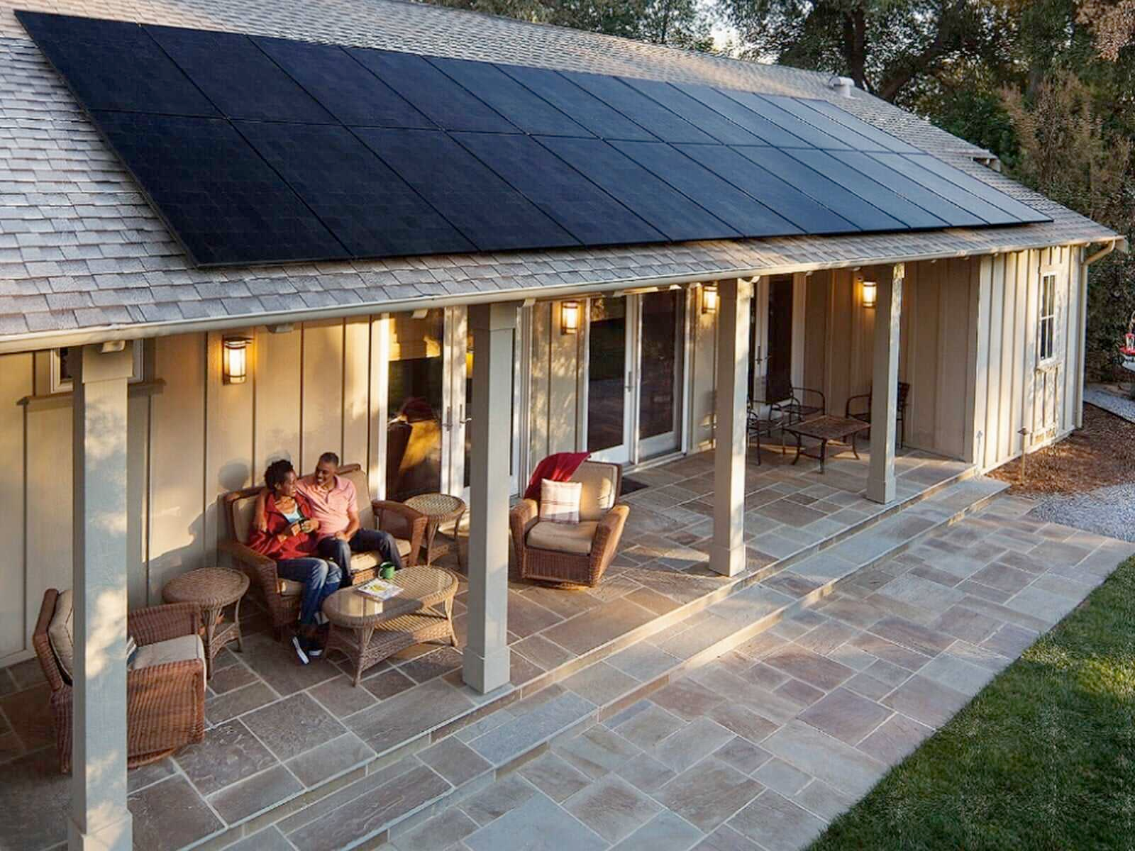 Two people are sitting on a porch of a house with solar panels on the roof.