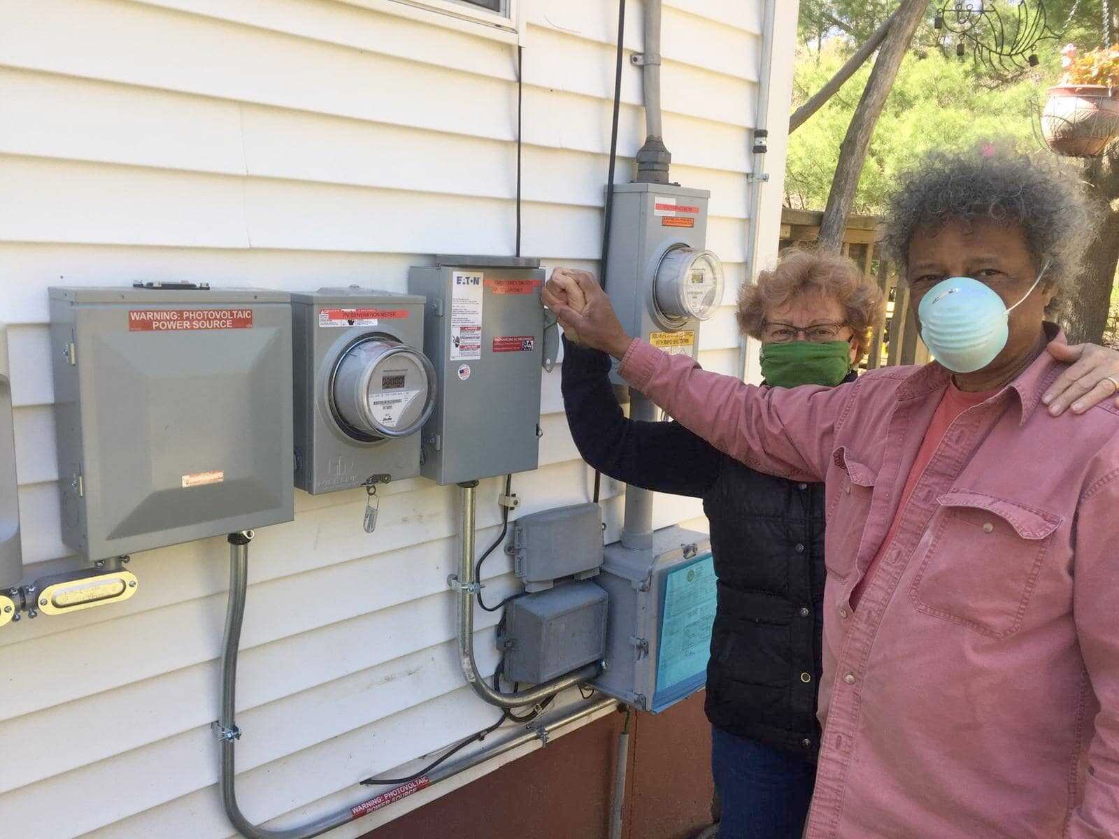 A man and a woman wearing masks are pointing at electrical boxes on the side of a house.