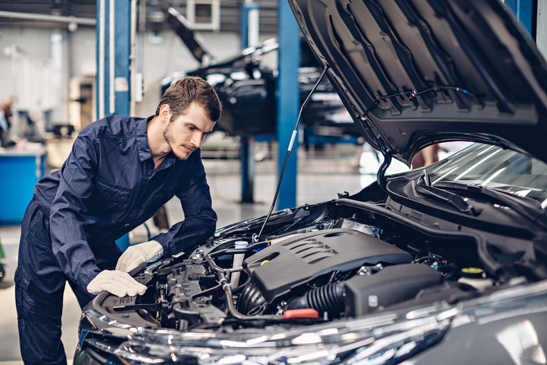 A mechanic in a blue uniform repairs a car, focused on the engine with tools in hand.