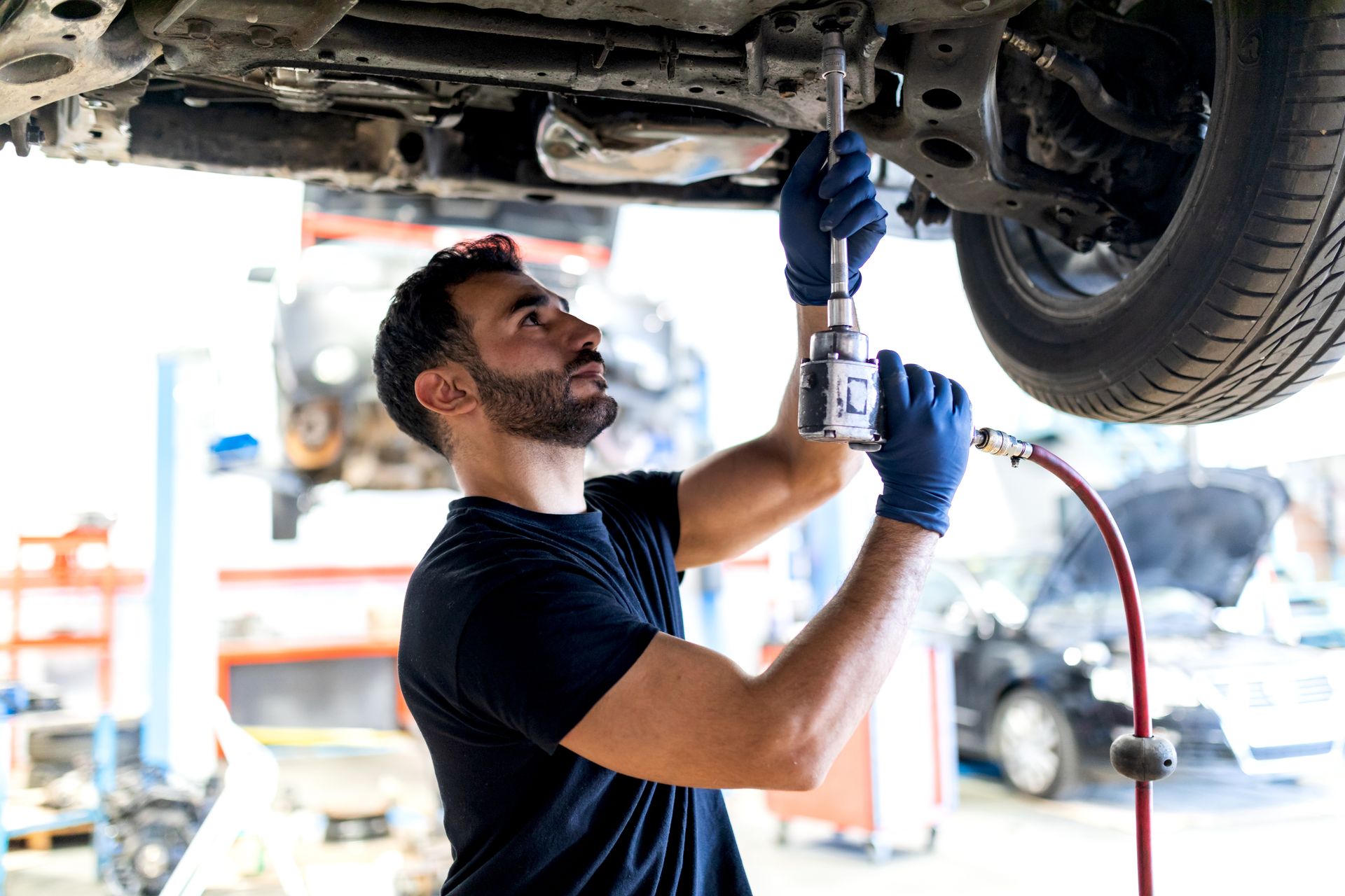 Photograph of a mechanic using a power tool to work on the underside of a lifted vehicle in an auto