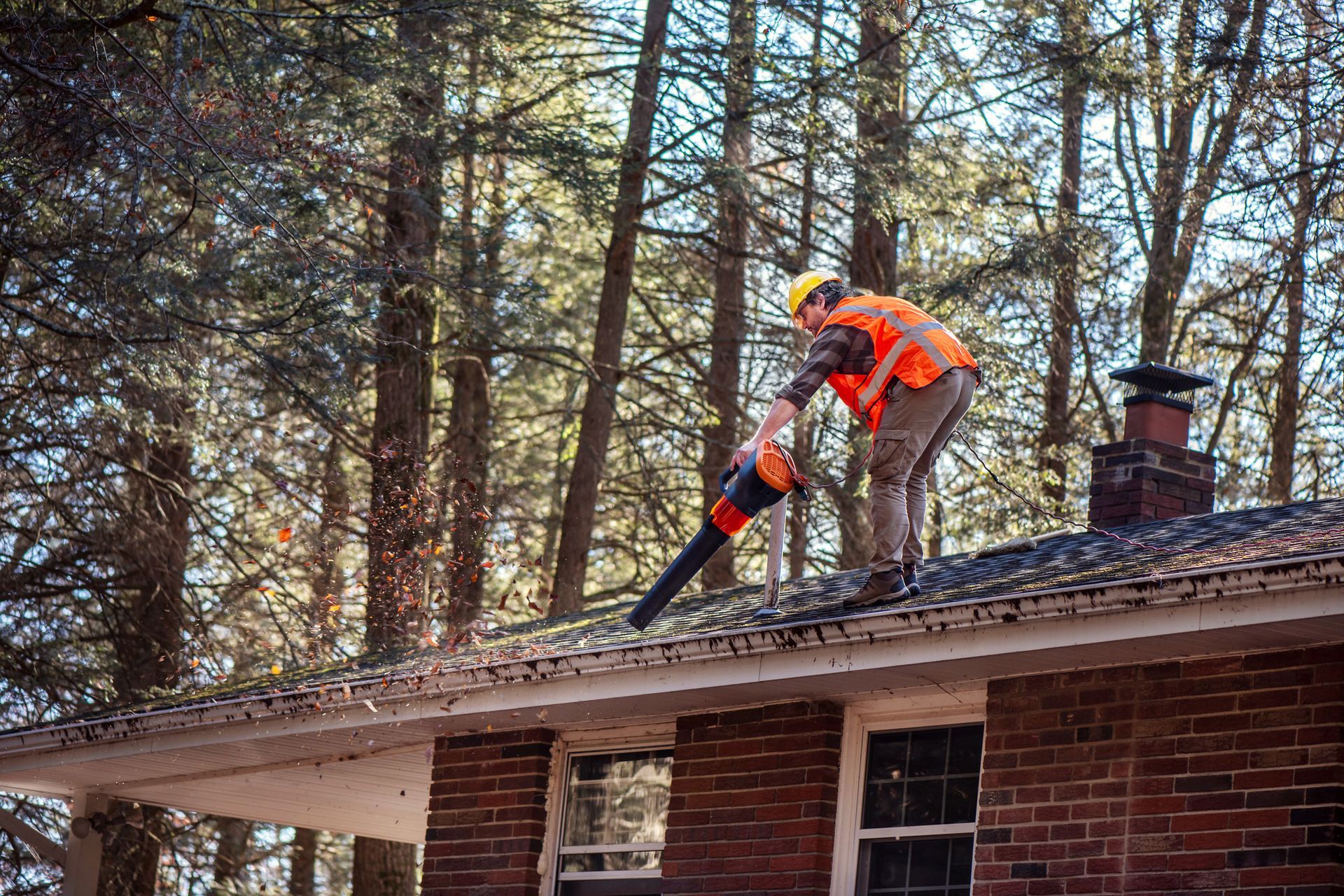A male contractor in a safety vest uses a leaf blower to clean debris from the shingle roof