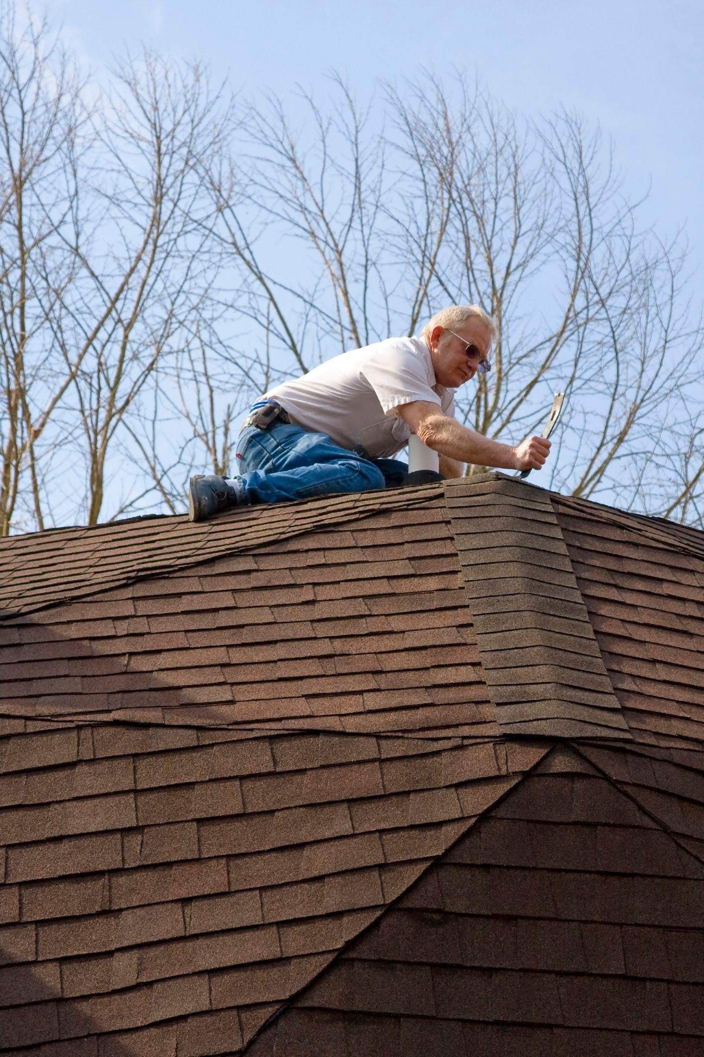 Roof contractor sits on top of, and checks shingled roof with blue sky as background.