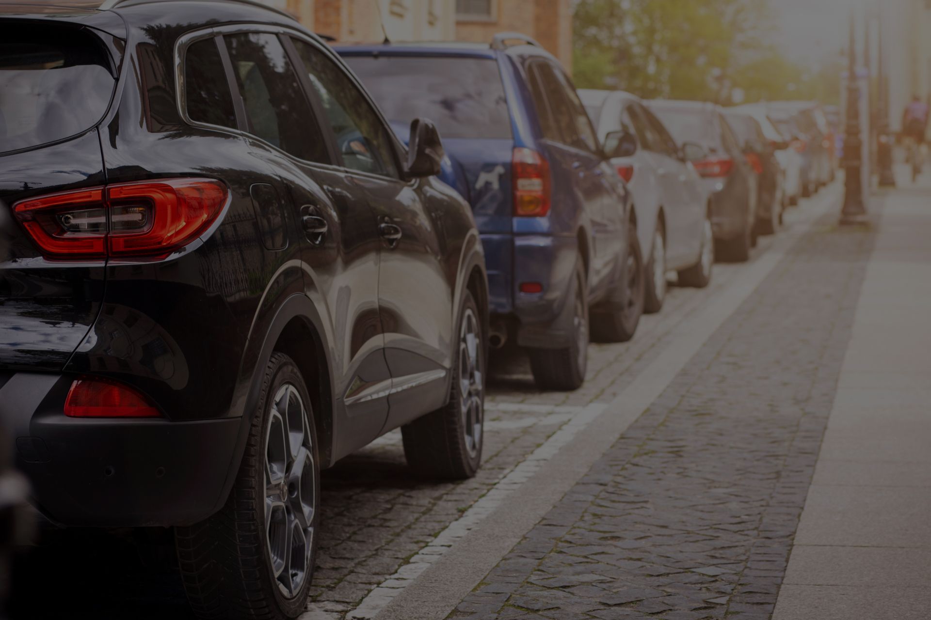 Cars parked along a cobblestone street. Black SUV in focus, other cars fading in background. | Importspor