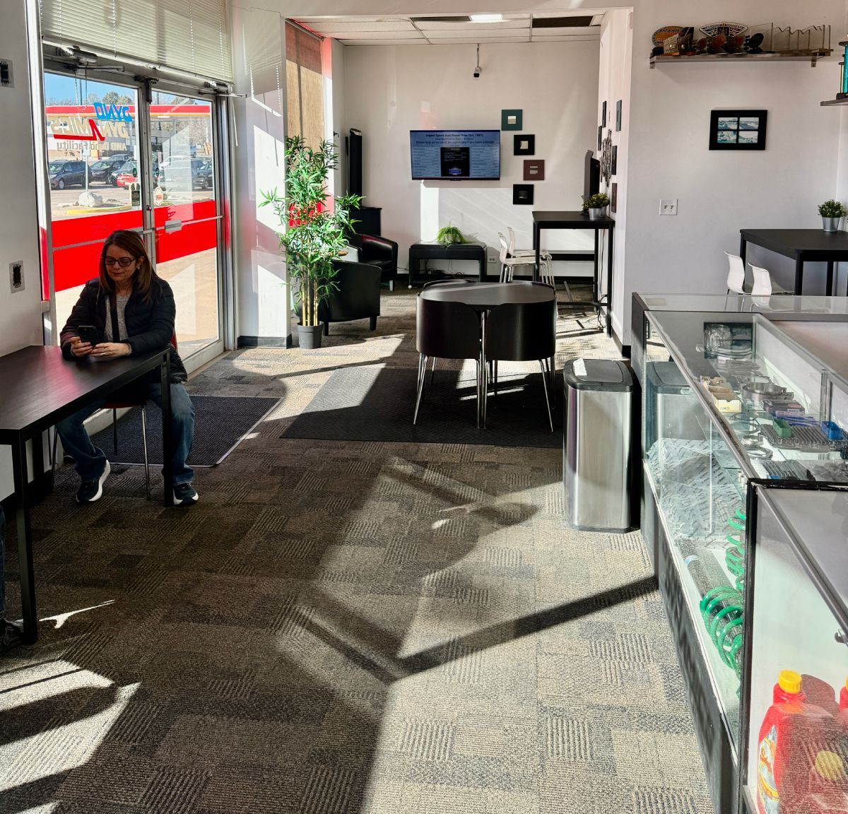 Waiting area with black couches, table, TV, plants, and vending machine. Sunlight streams in from a red-framed door. | Importsports