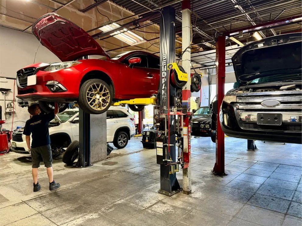 Mechanic working on a red car lifted on a hoist in an auto repair shop. Other vehicles visible. | Importsports