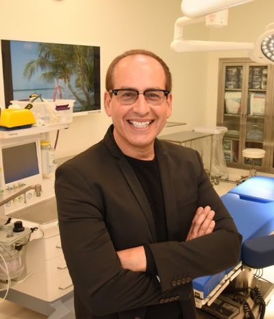 Smiling man in glasses and black blazer in medical room, arms crossed.