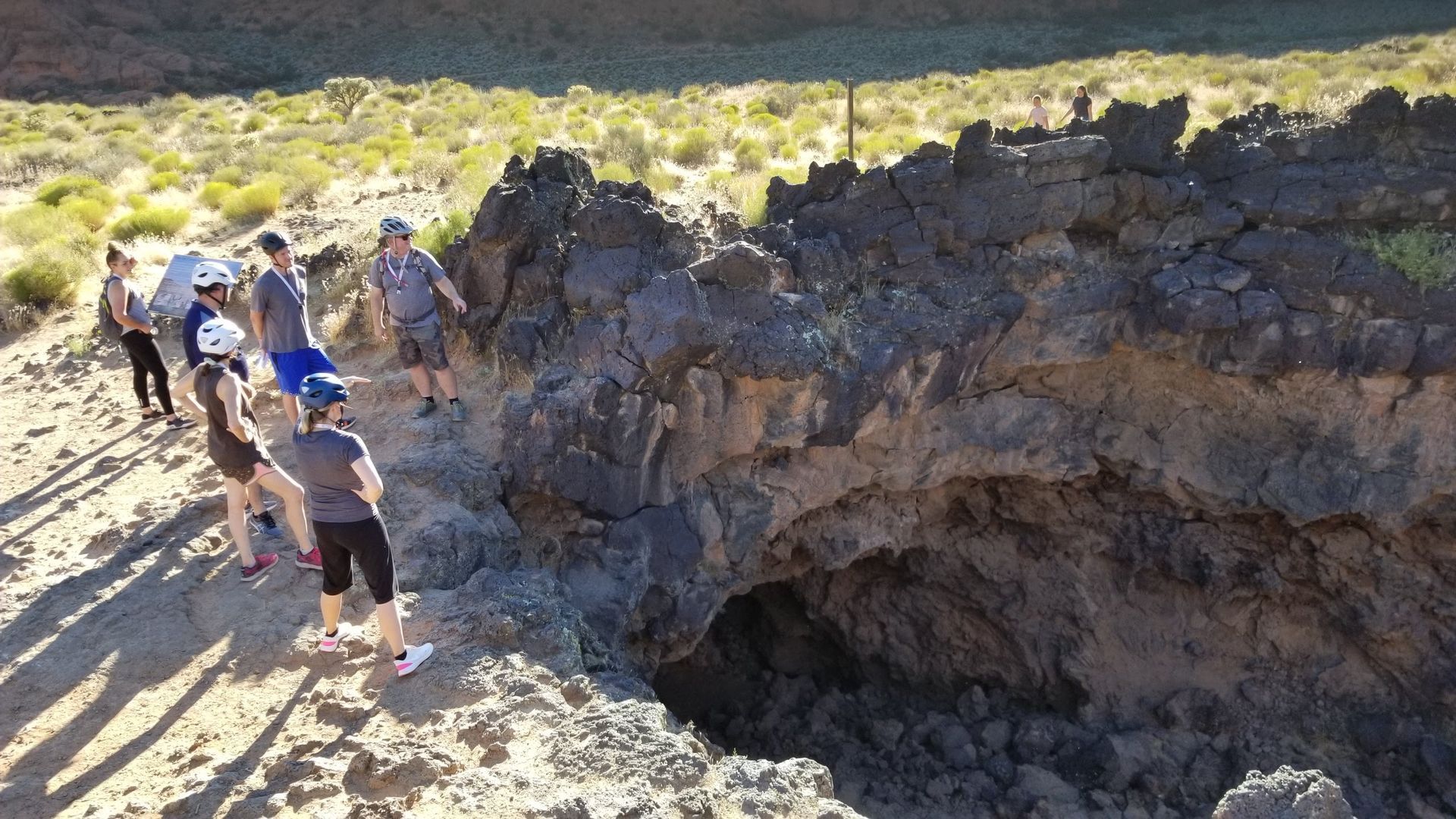 A group of people are standing in front of a cave formed by lava flow in Snow Canyon.