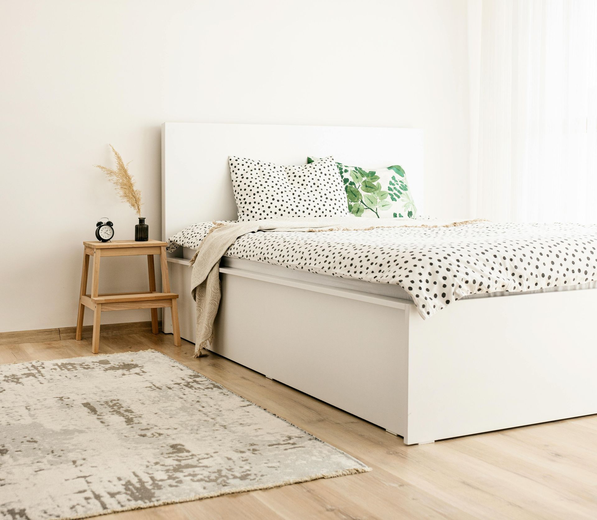 White bed with patterned bedding, wood floor, rug, and side table with decor.