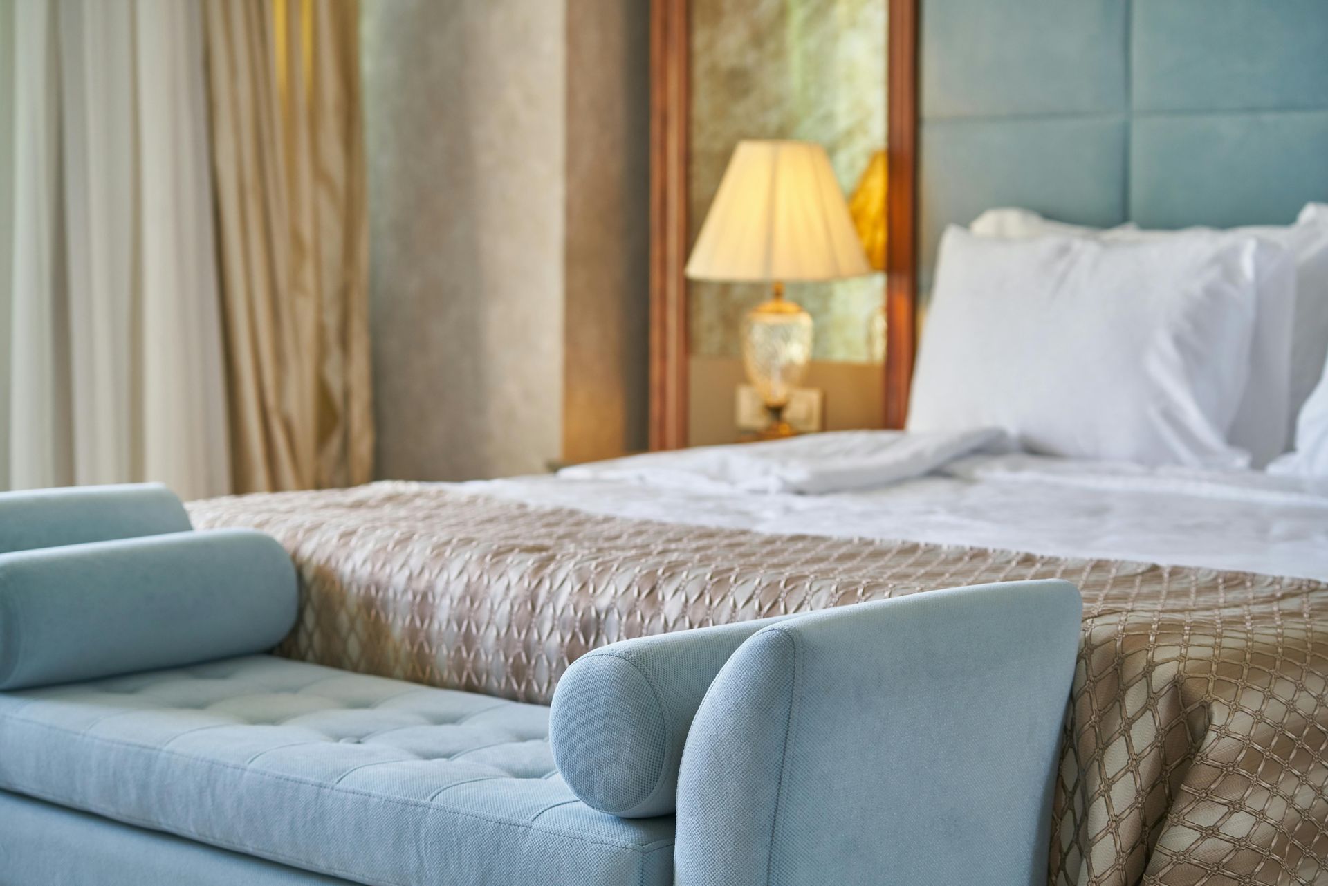 Bedroom with a light blue bench, bed with white and beige linens, and a lit lamp.