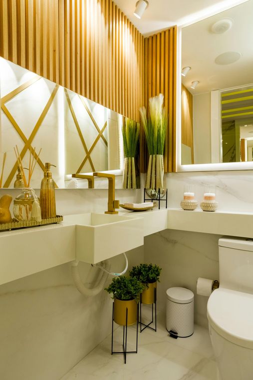 Modern bathroom with wooden accent wall, gold fixtures, white sink and toilet, and potted plants.