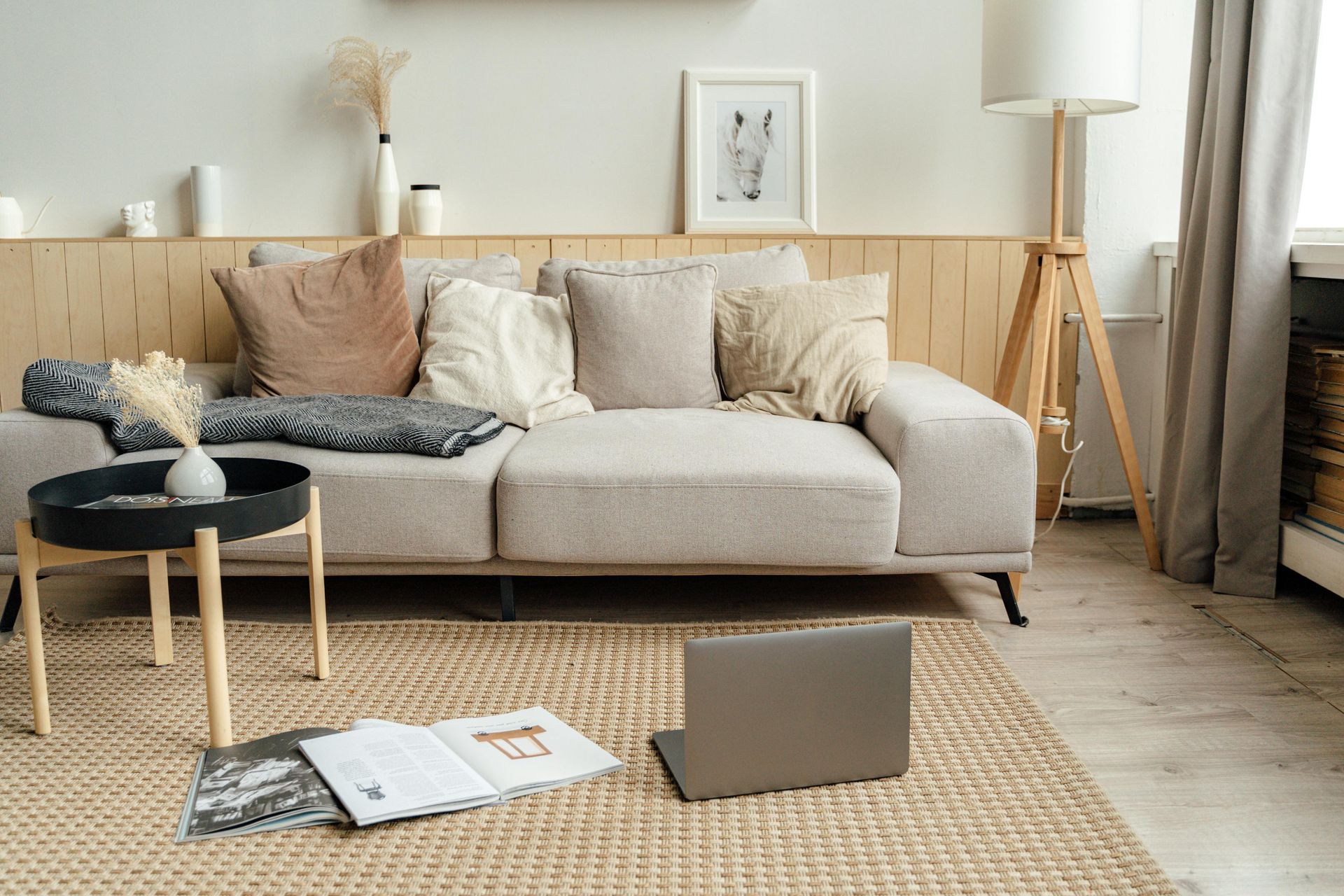 Beige couch with pillows, lamp, coffee table, laptop, and rug in a bright living room.