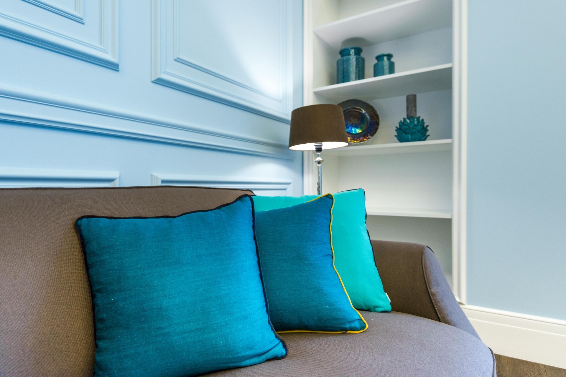 Brown sofa with three teal pillows, lamp, and white shelves with turquoise vases.