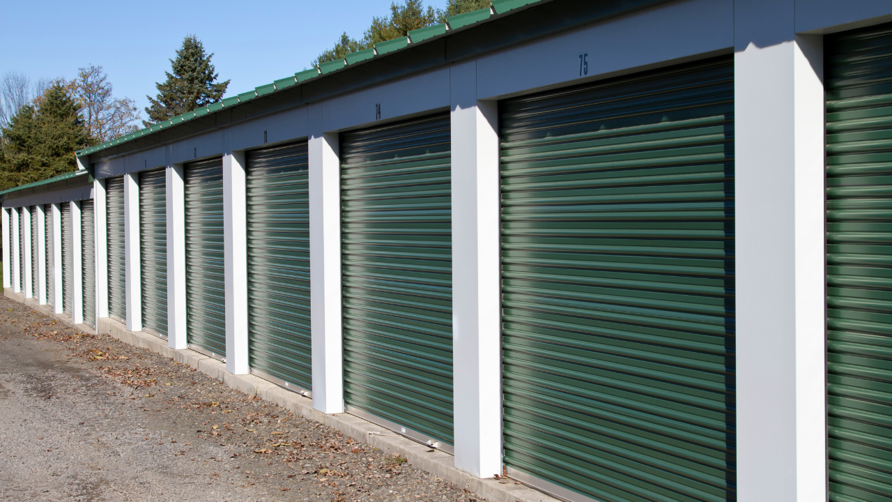 Row of green storage units with white frames and numbered doors, outdoors.